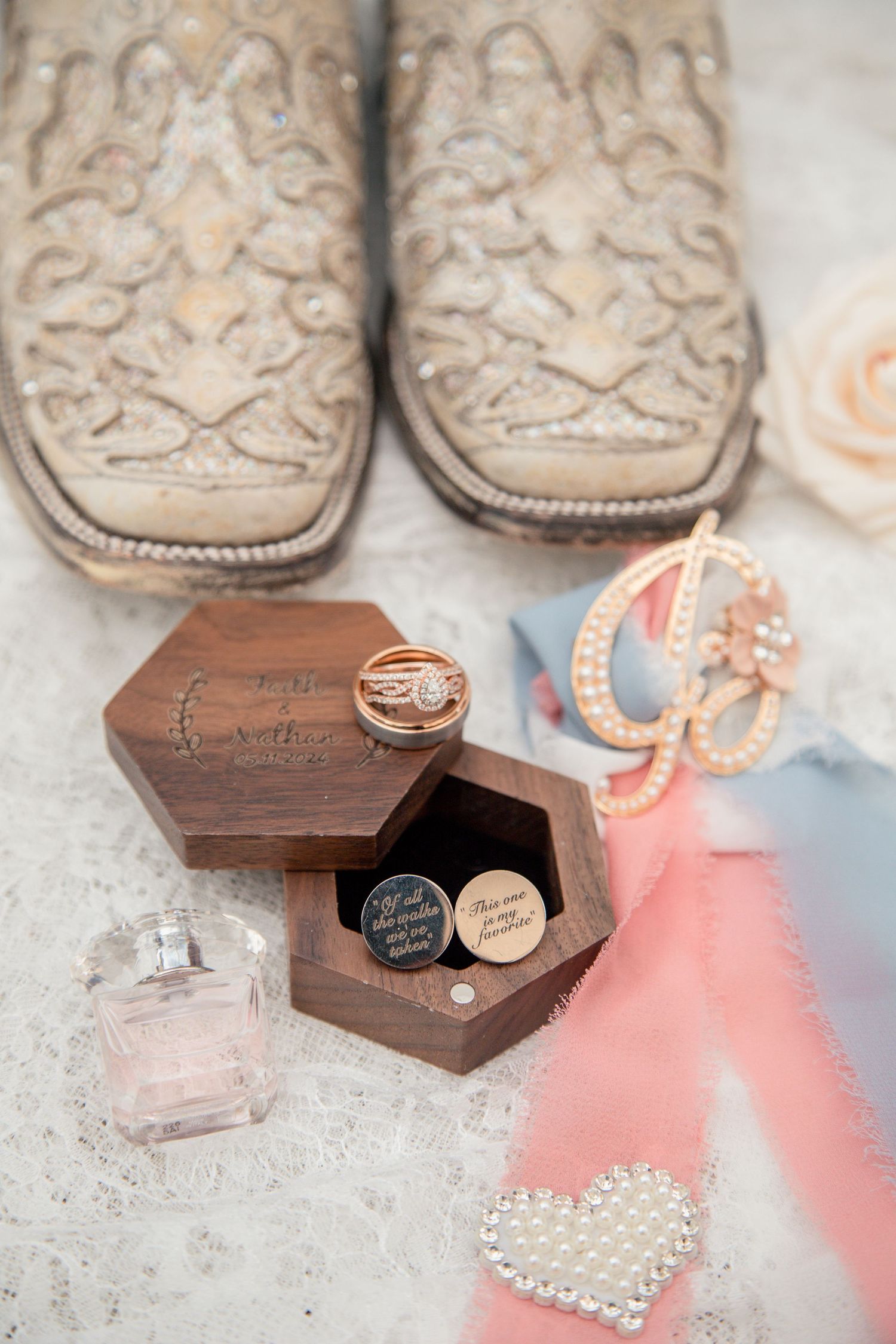 Wedding flat lay featuring cowboy boots, wooden ring box with gold bands, perfume, monogram brooch, and pink ribbon details.