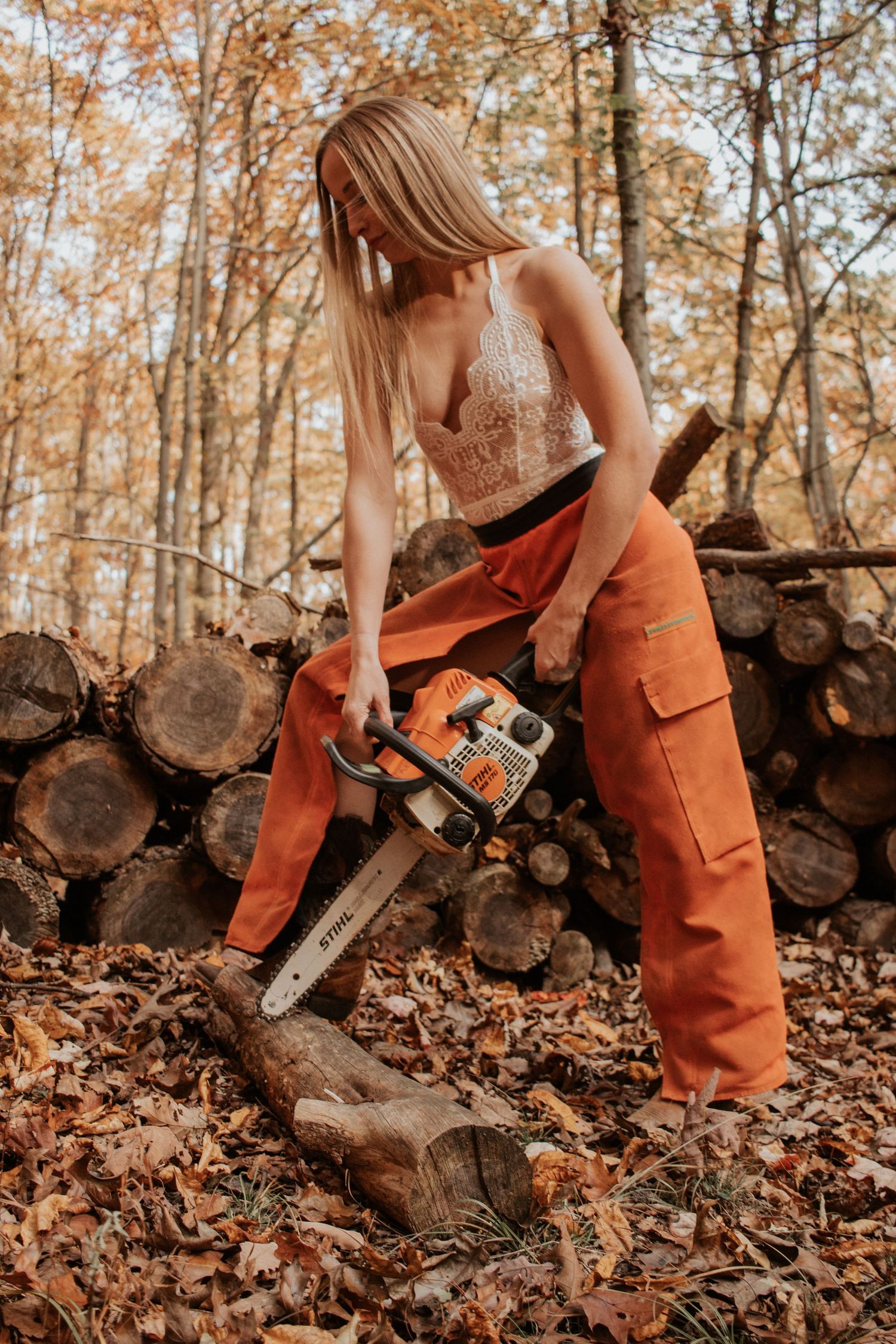 Blonde woman in orange cargo pants using a chainsaw to cut wood in an autumn forest.