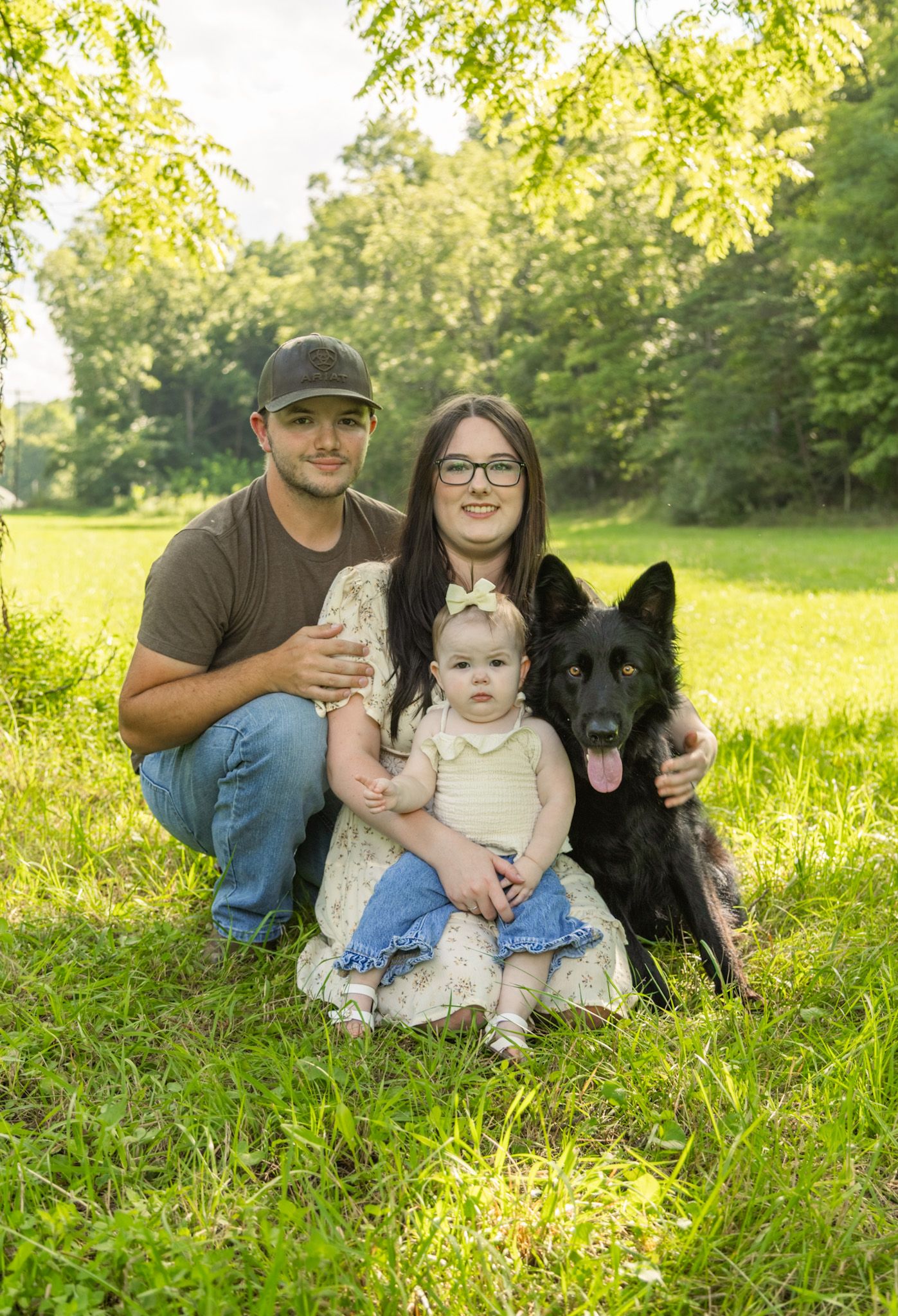 Smiling family of three with a black German Shepherd posing together on a sunny grassy lawn surrounded by green trees.