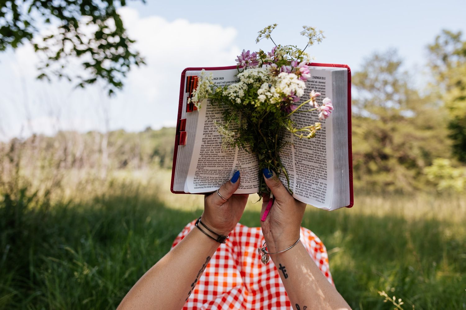 Woman holding open a red Bible with colorful wildflowers bouquet against a sunny green meadow background.