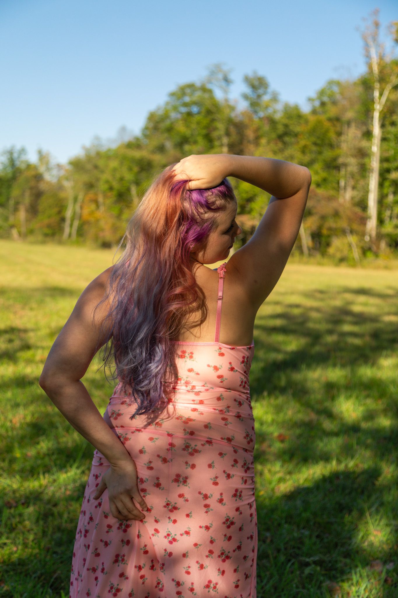 Woman in pink floral sundress with purple hair poses in sunny green meadow with trees in background.