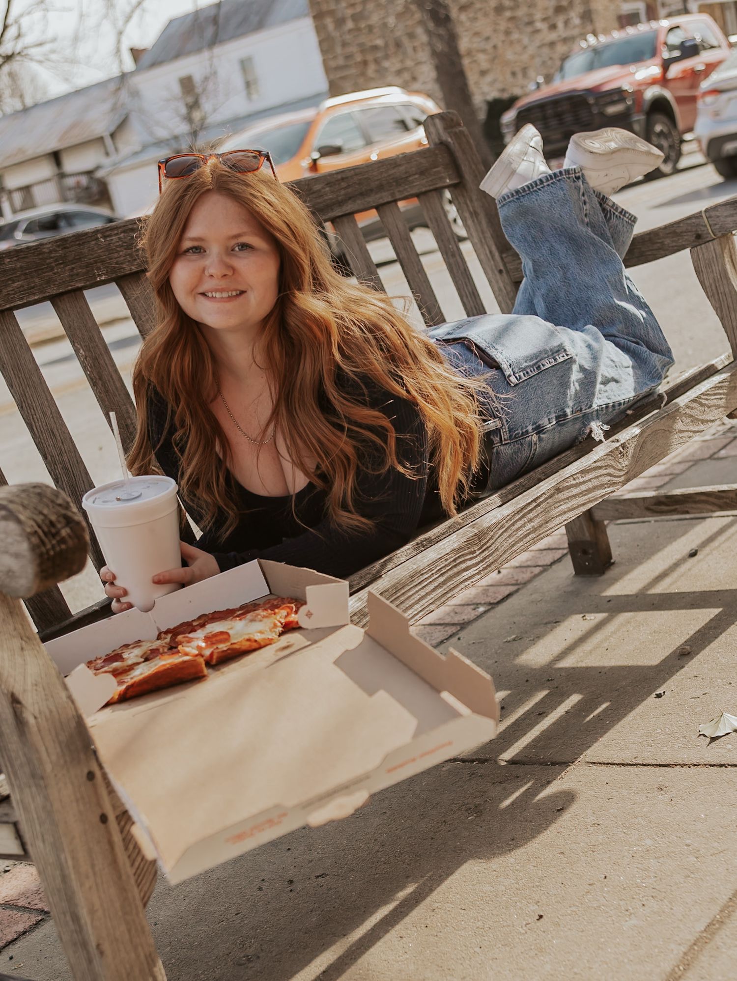 Smiling redhead girl lying on a wooden bench outdoors with pizza and a drink on a sunny day.