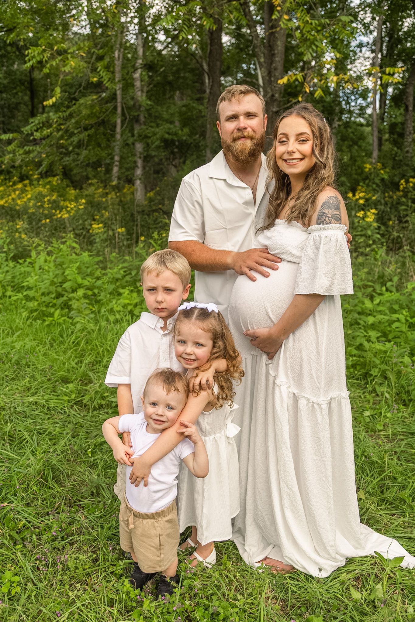 Expecting couple in white outfits pose with three young children in a green wooded meadow for an outdoor maternity family photo.