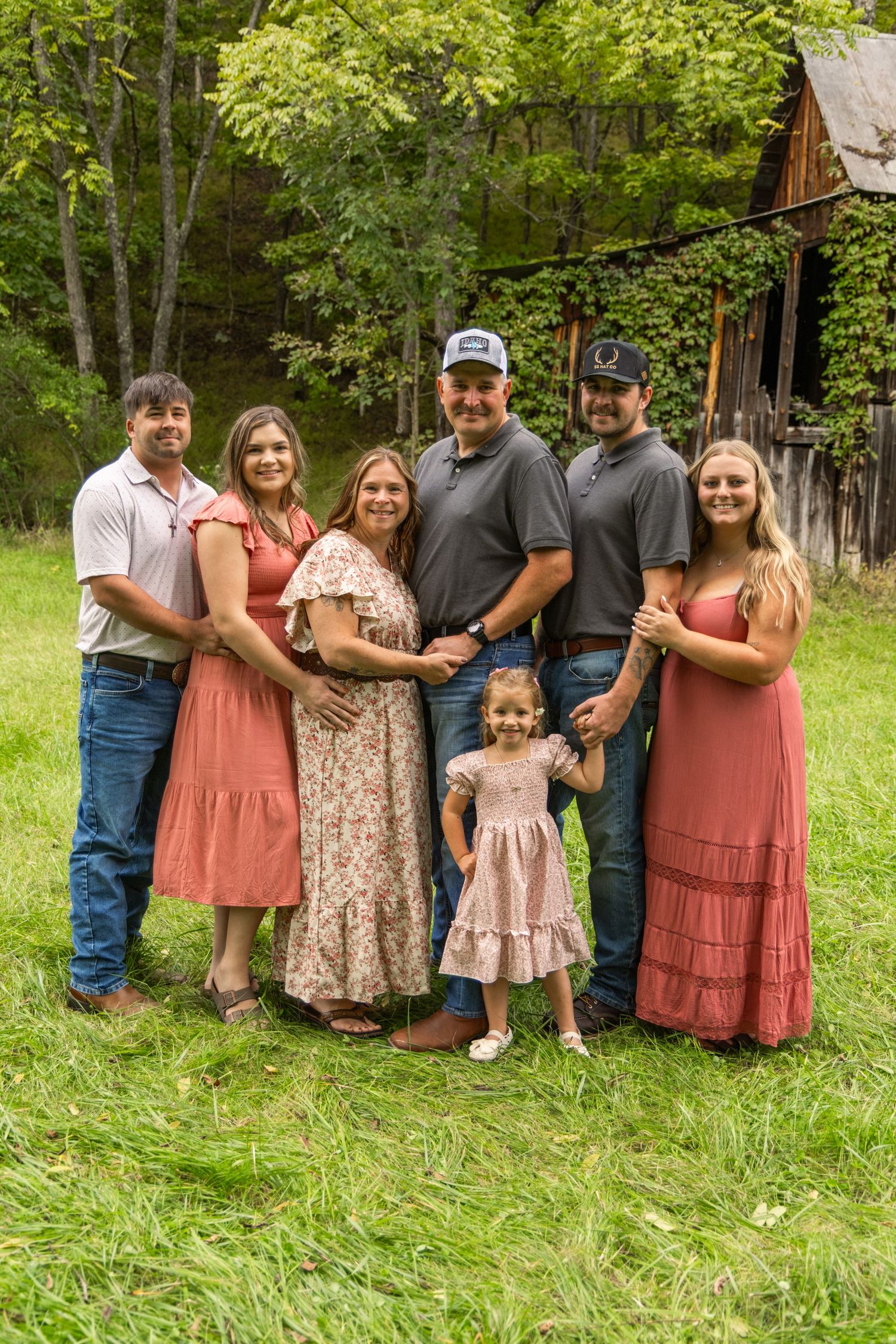 Family of seven in coordinated coral and neutral outfits posing together on a green lawn in front of a rustic barn and trees.