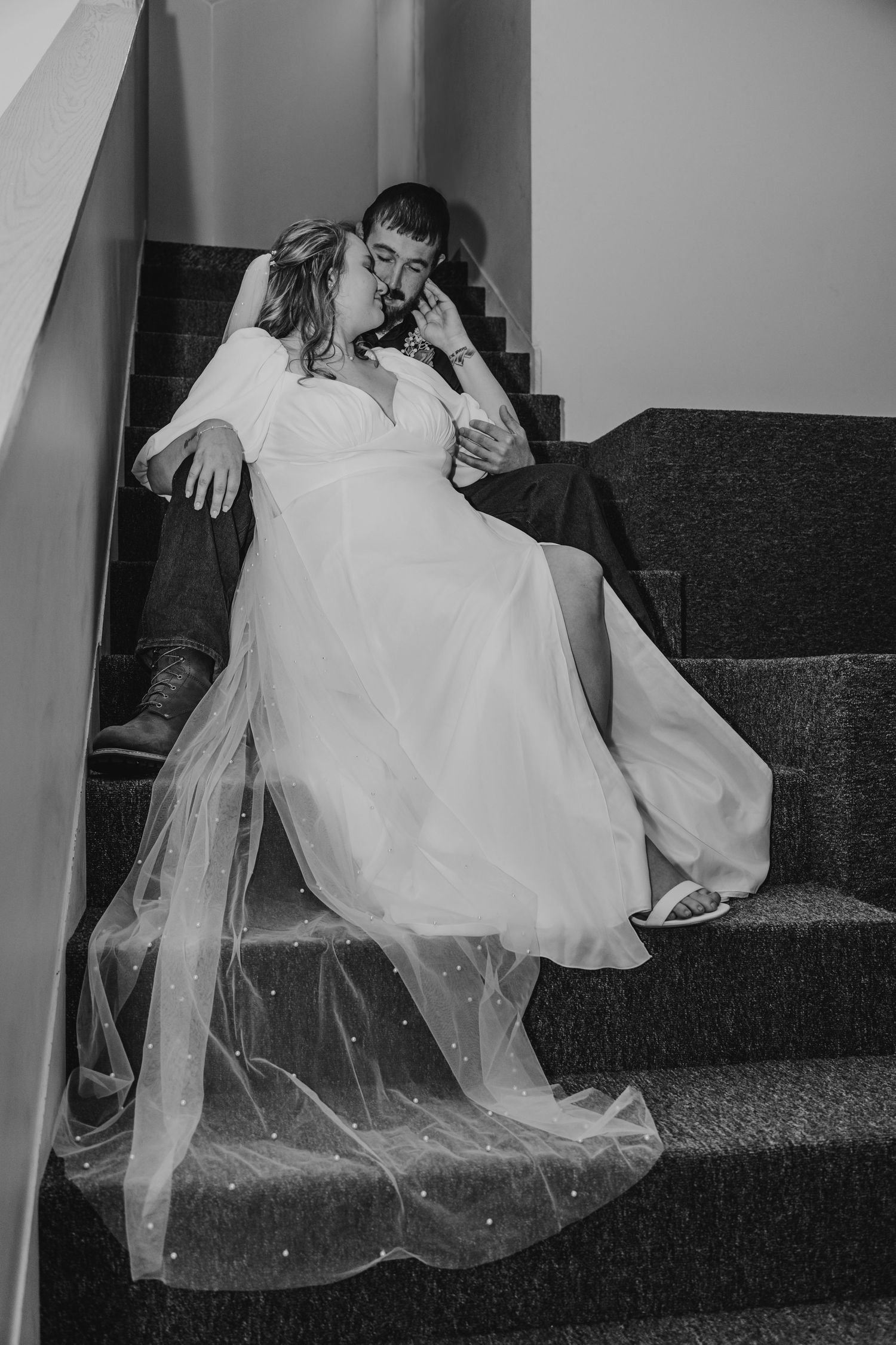 Black and white romantic photo of a bride and groom sitting closely together on carpeted stairs.