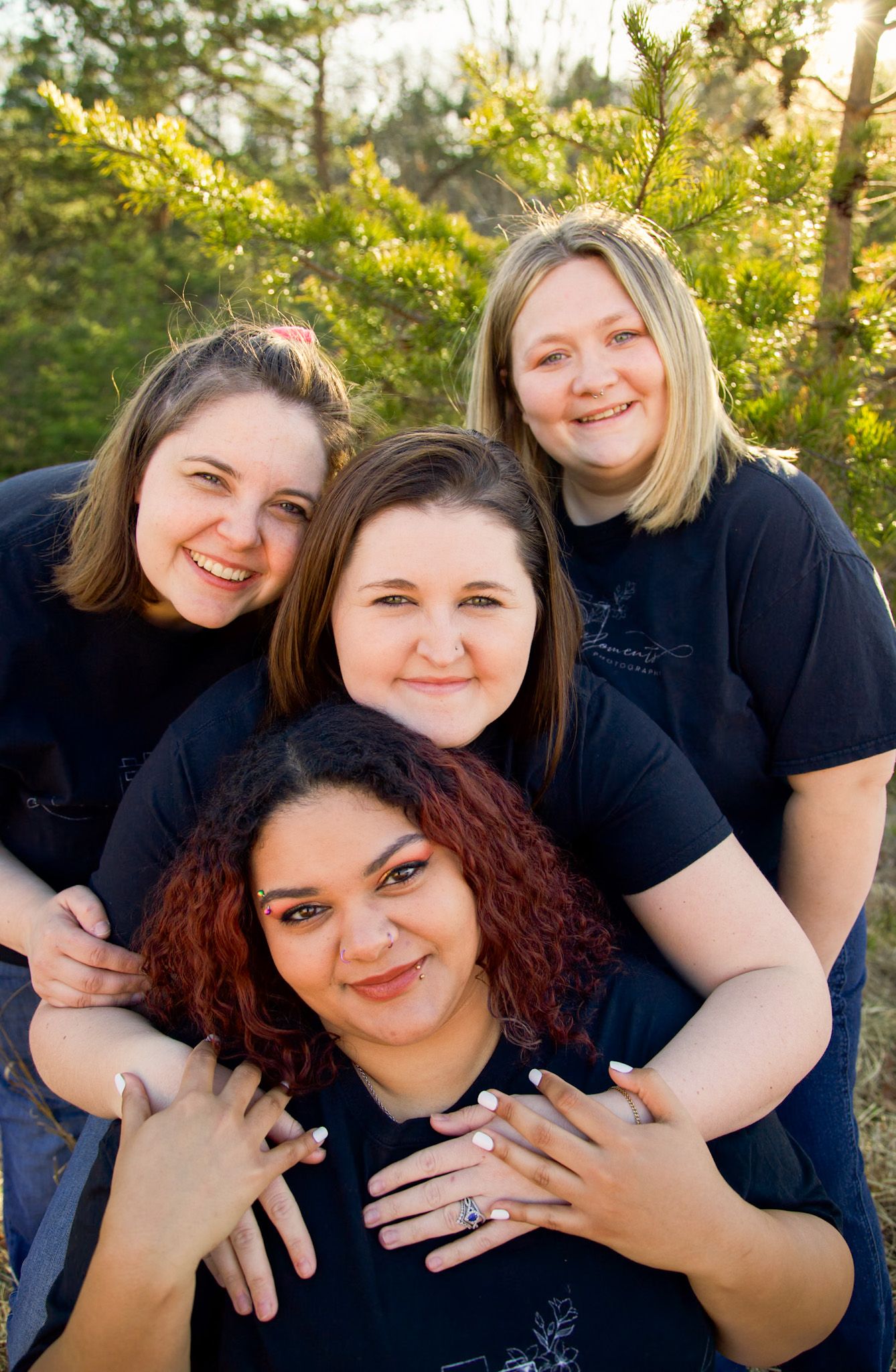 Four women in black t-shirts smiling closely together outdoors in warm golden sunlight near trees.