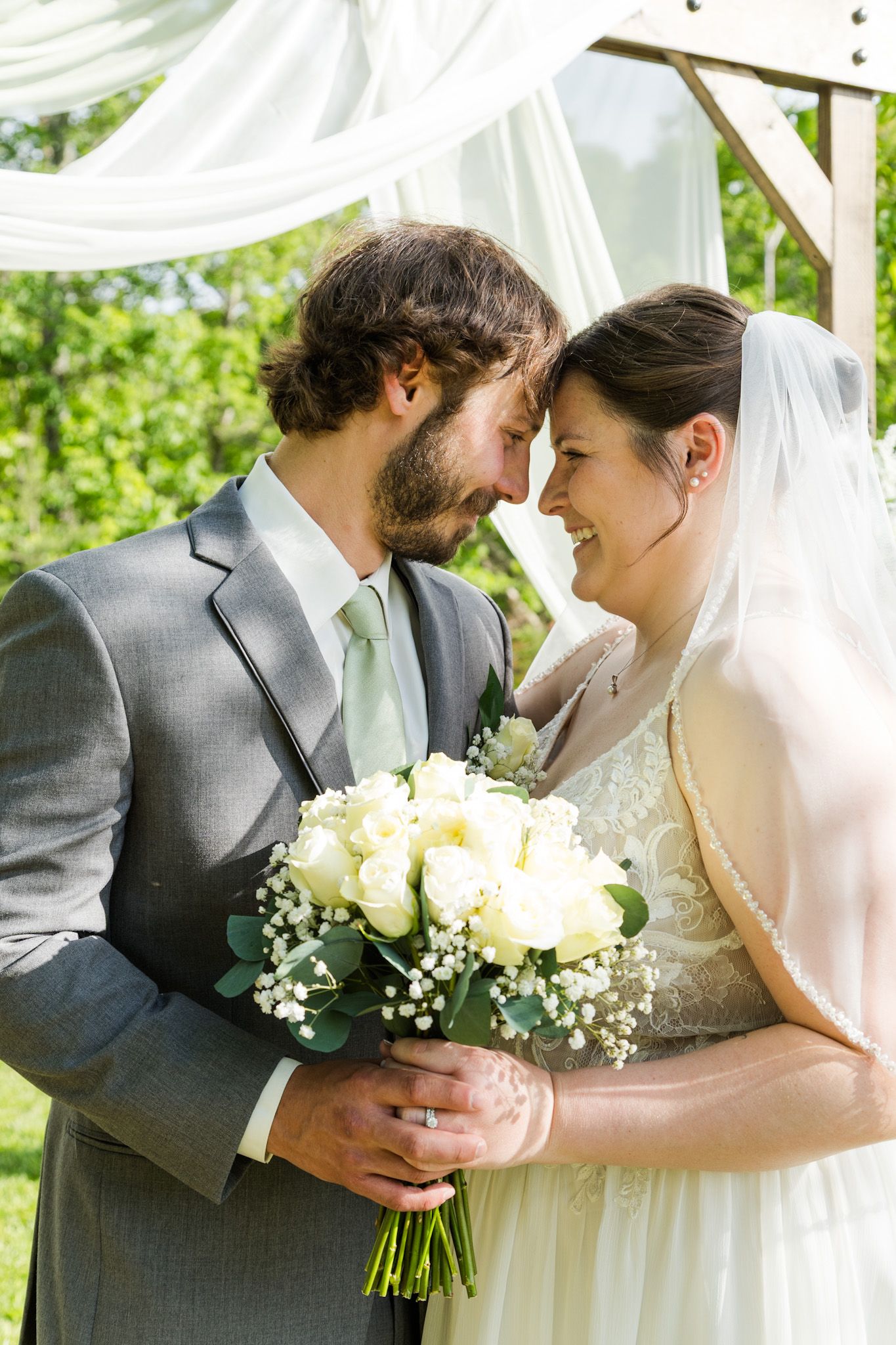 Bride and groom touching foreheads and smiling while holding white rose bouquet at outdoor wedding ceremony.
