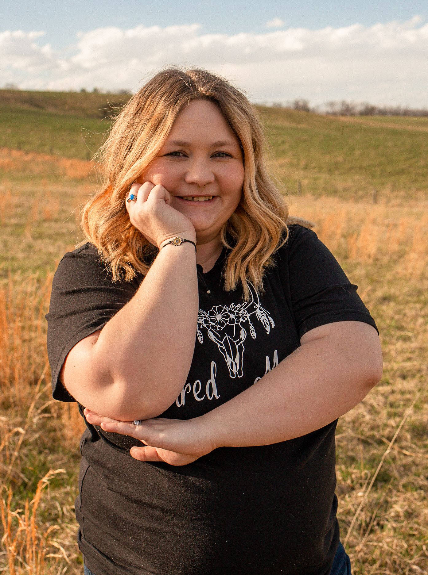 Cheerful blonde woman in a black Treasured Moments shirt resting her chin on her hand in a golden field.
