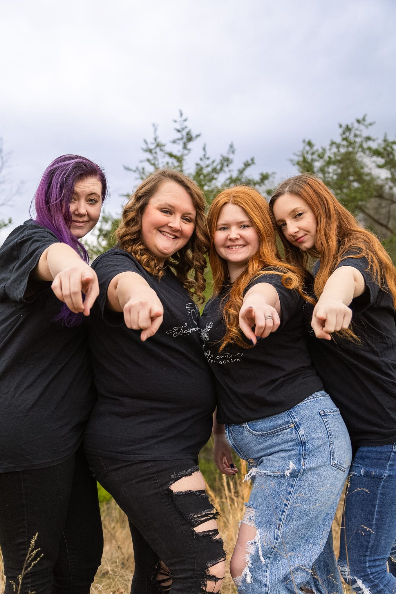 Four young women in black shirts pointing at camera outdoors.