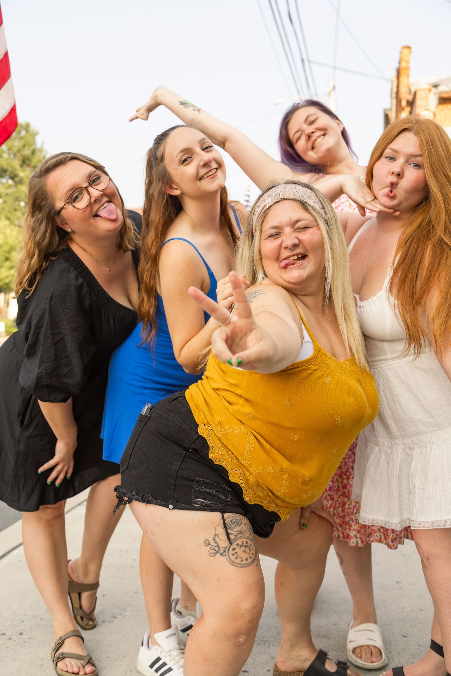 Five young women posing playfully outdoors, making fun faces and peace signs near an American flag.