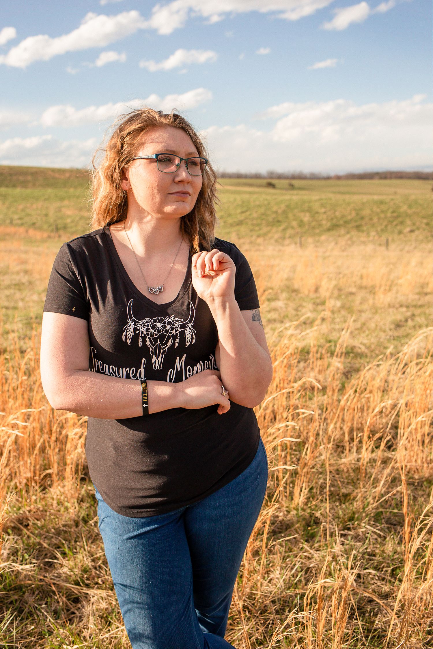 Woman with glasses in a black Treasured Moments shirt standing in a golden field under a partly cloudy sky.
