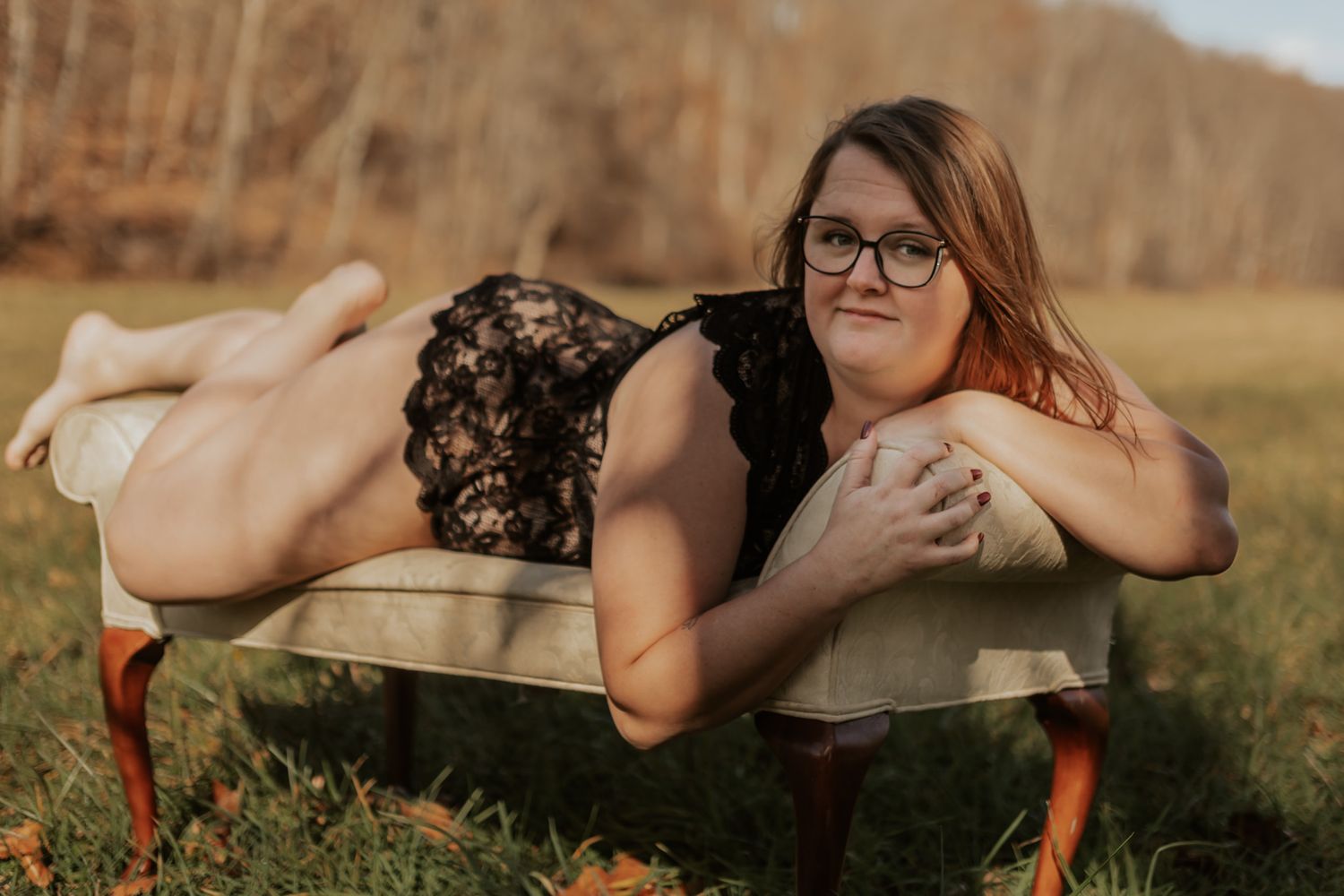 Woman in black lace bodysuit lies on vintage settee in outdoor autumn field setting.