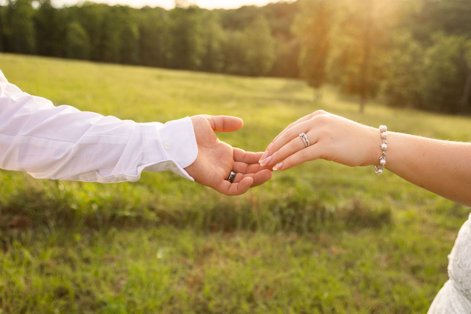 Bride and groom gently touching hands outdoors in a sunlit green field, bride wearing diamond ring and pearl bracelet.