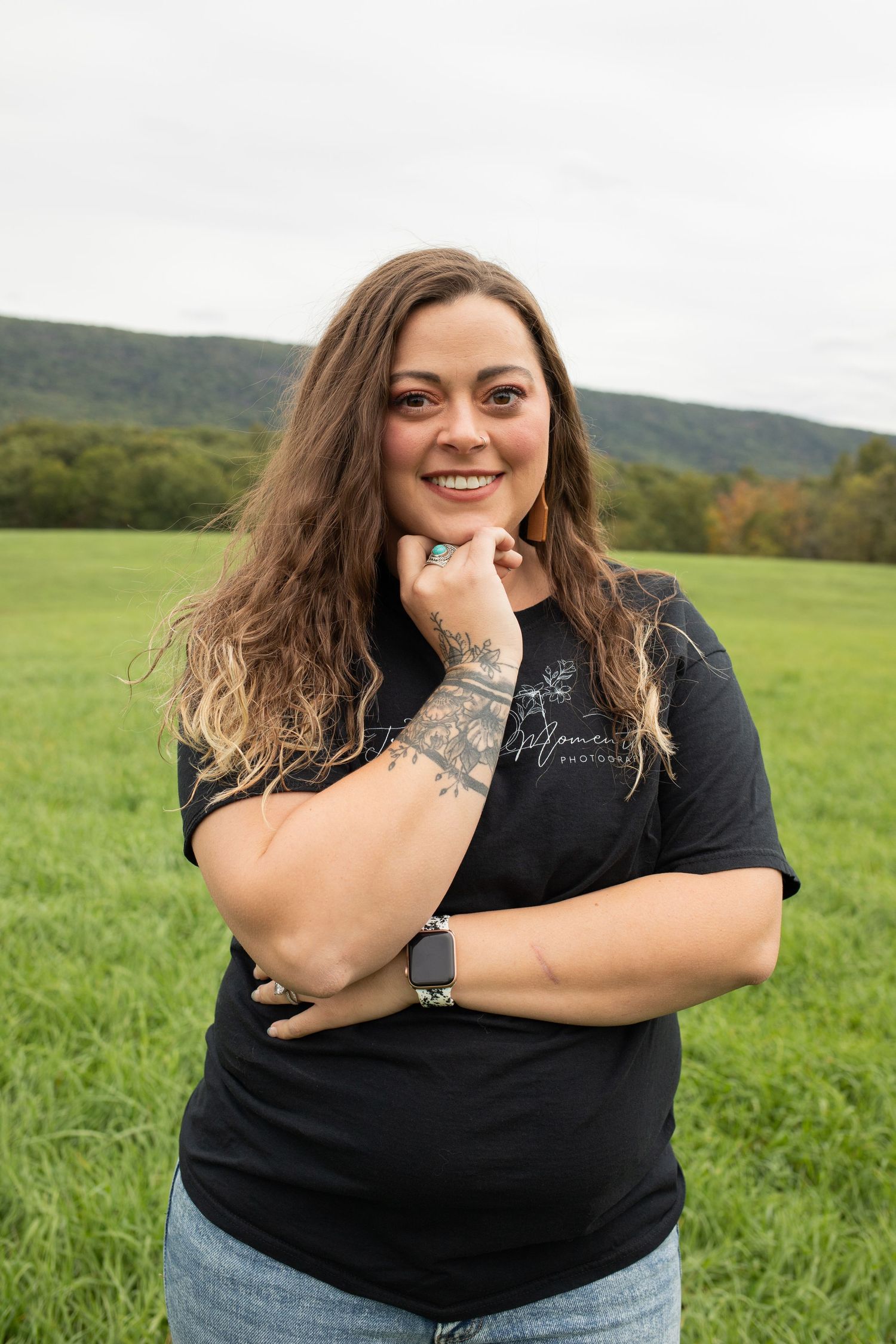 Tattooed woman in black tee smiling in lush green meadow outdoors.