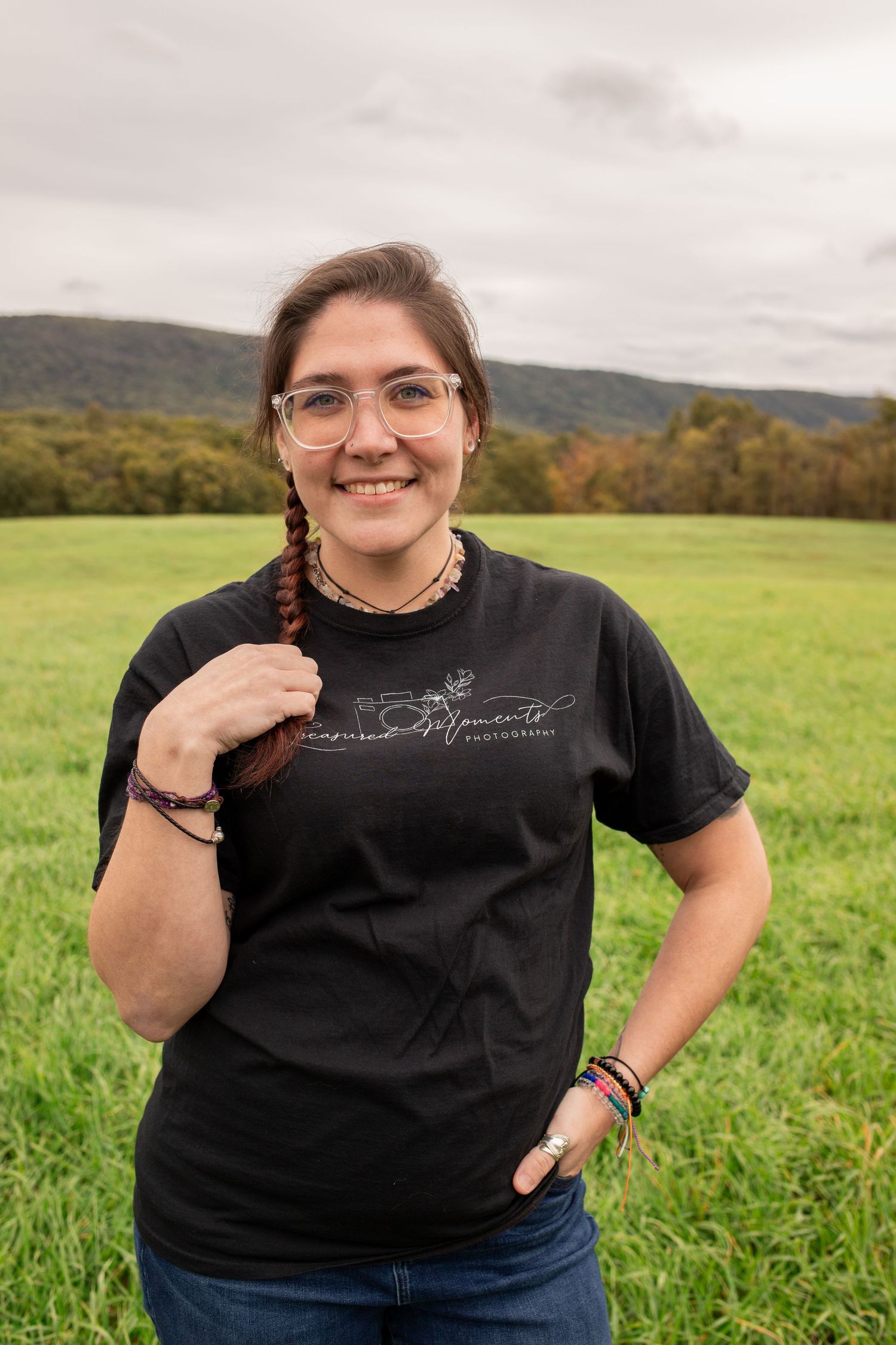 Smiling woman with glasses and braid in black tee in green field.
