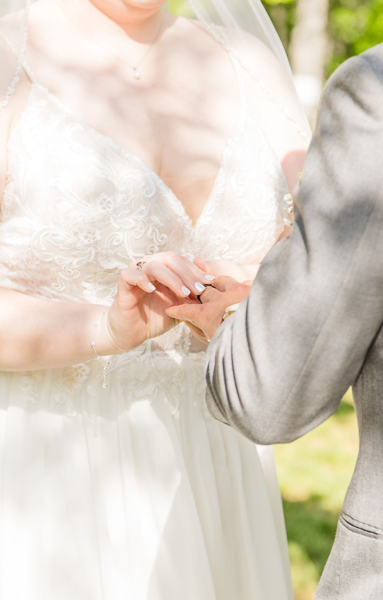 Bride in lace gown placing wedding ring on grooms finger during outdoor ceremony, soft sunlight filtering through veil.
