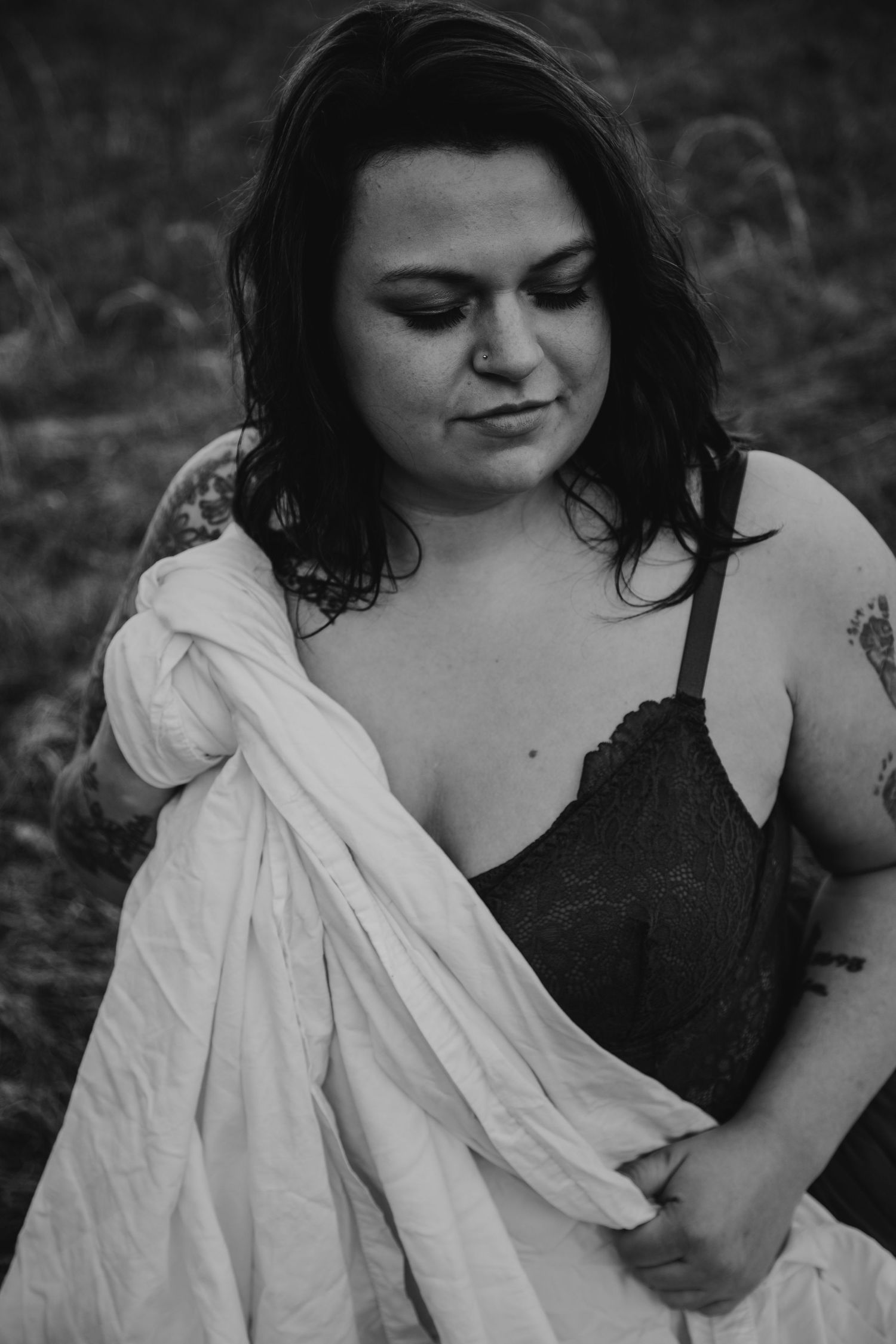 Black and white portrait of a tattooed woman in a black lace bralette holding a white cloth outdoors.