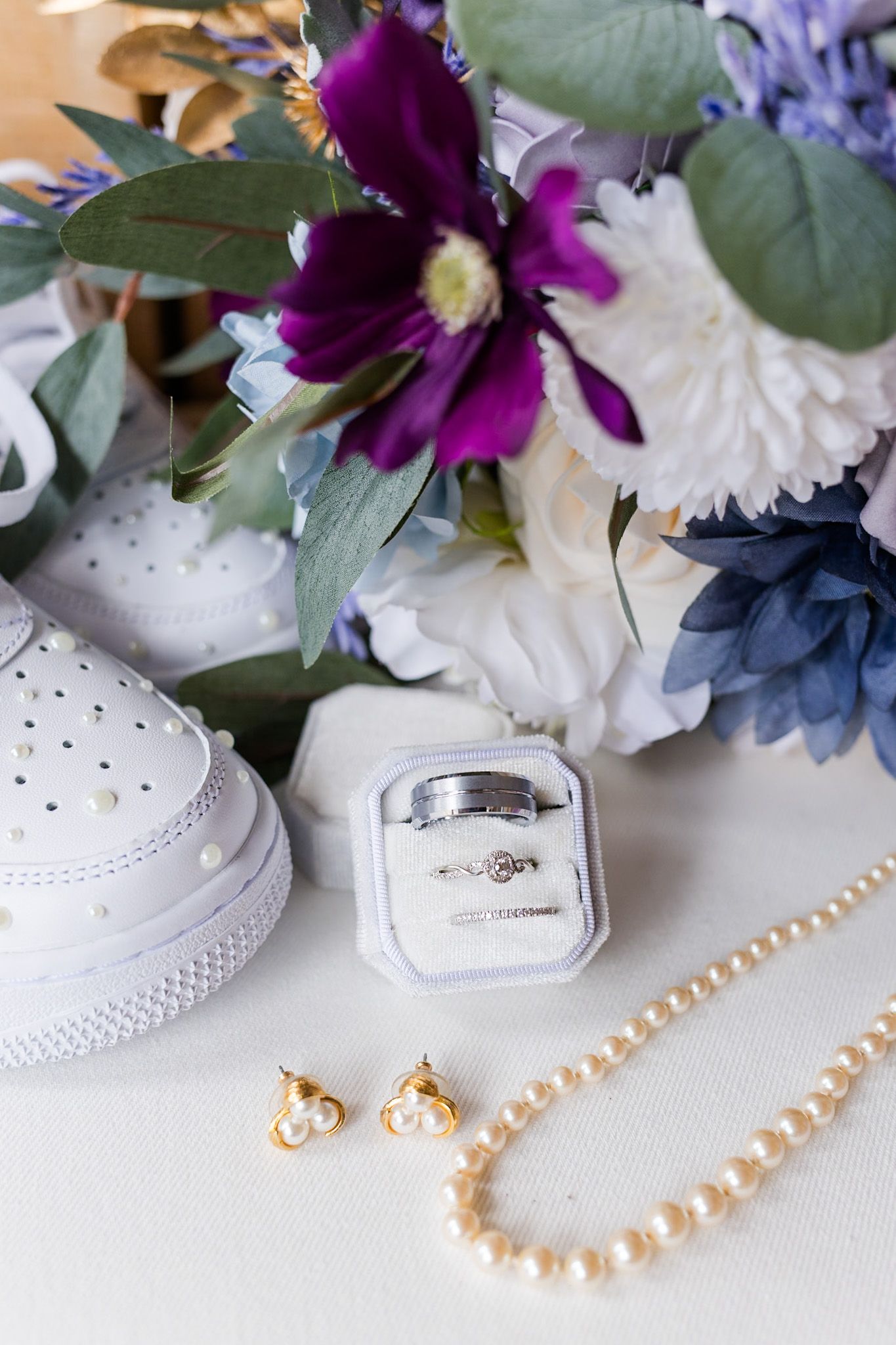 Wedding detail flat lay with white Nike sneakers, ring box, pearl necklace, earrings, and purple and white floral bouquet.