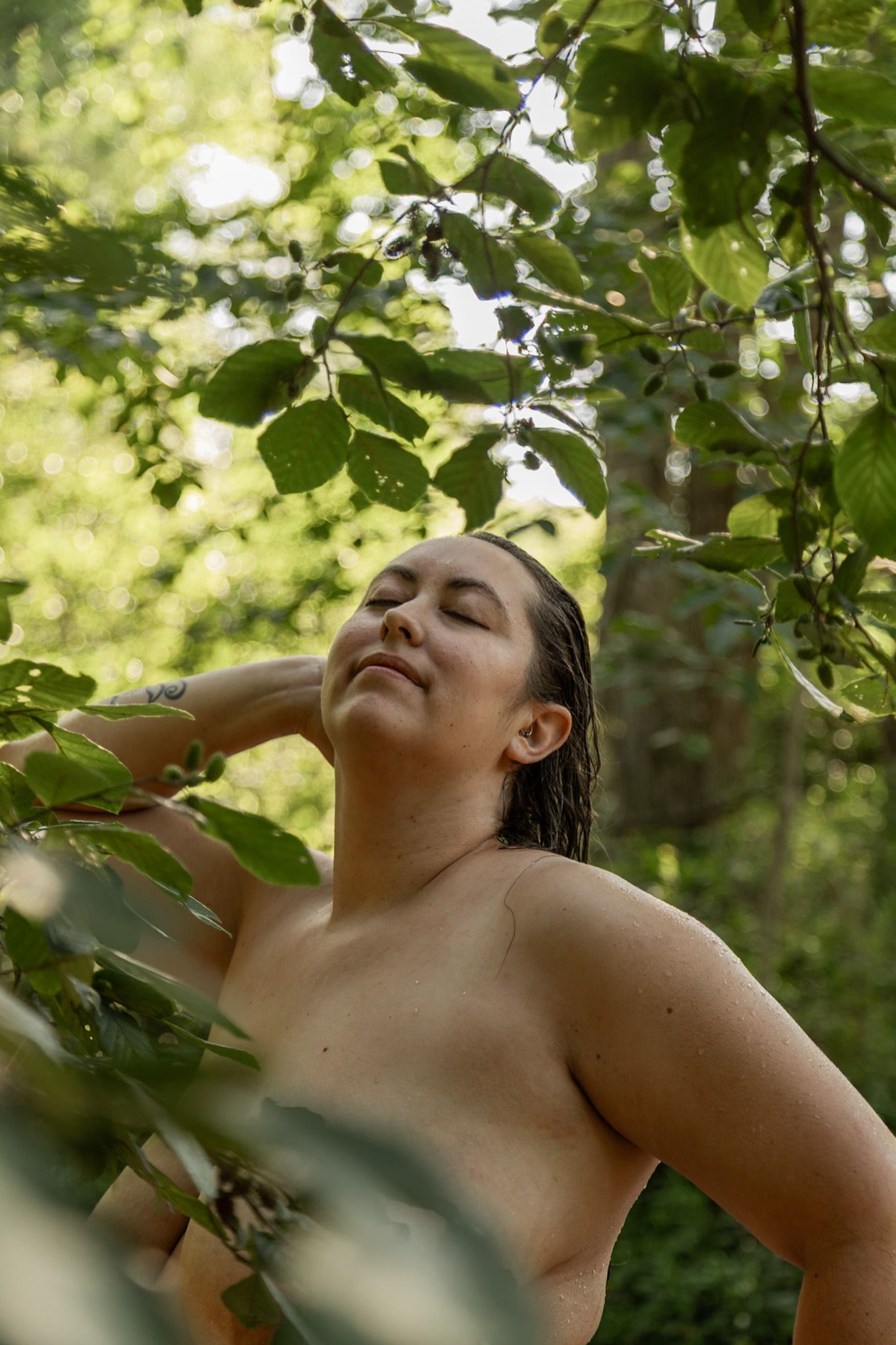 Woman with wet hair stands among green leaves in forest with eyes closed in peaceful pose.