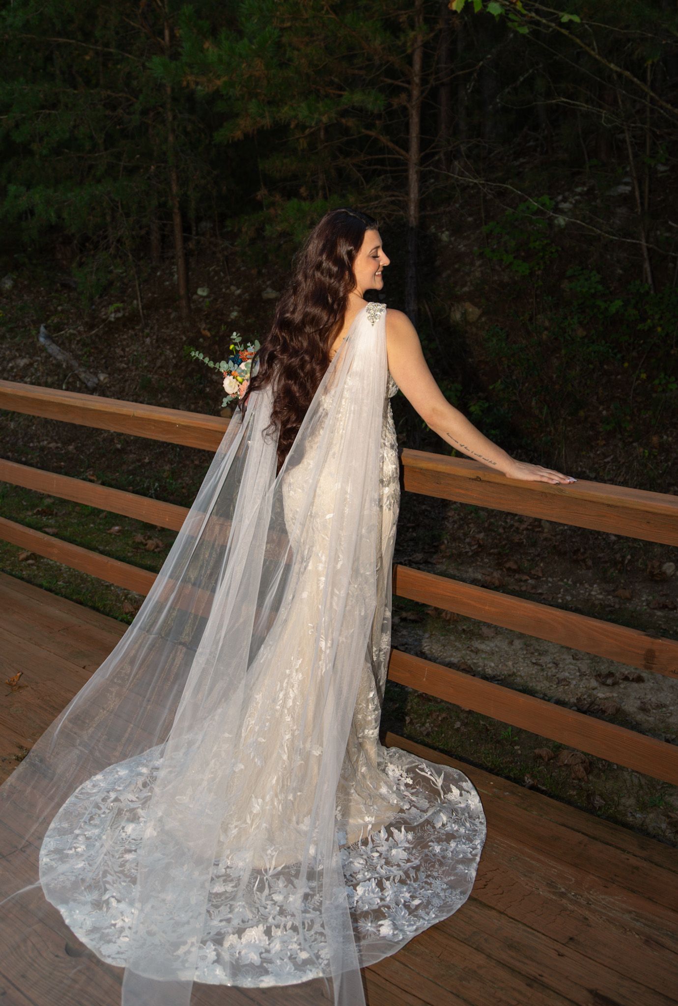 Bride in lace gown with long cathedral veil stands on wooden deck surrounded by forest.