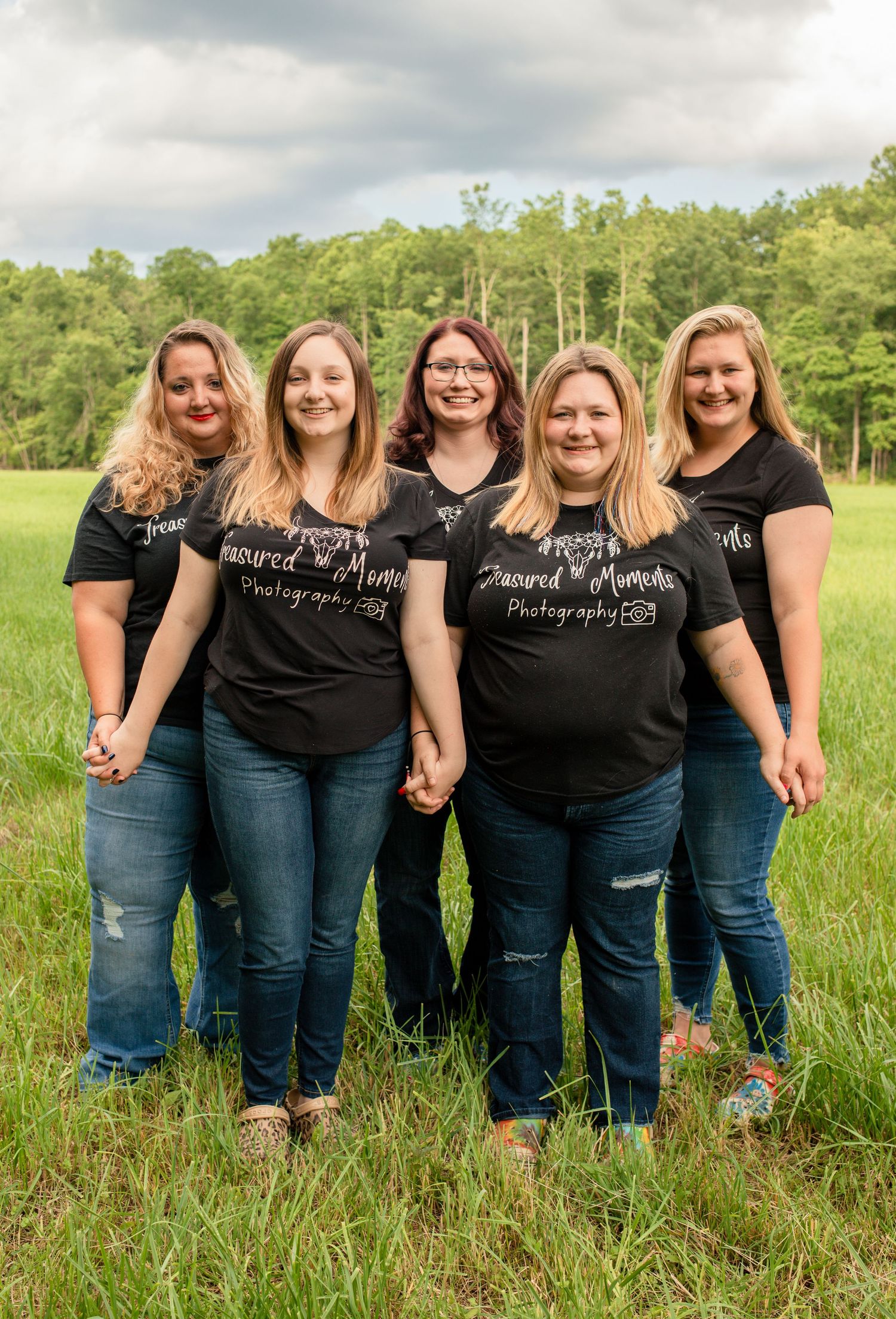 Five women in matching Treasured Moments Photography black t-shirts holding hands in green meadow.
