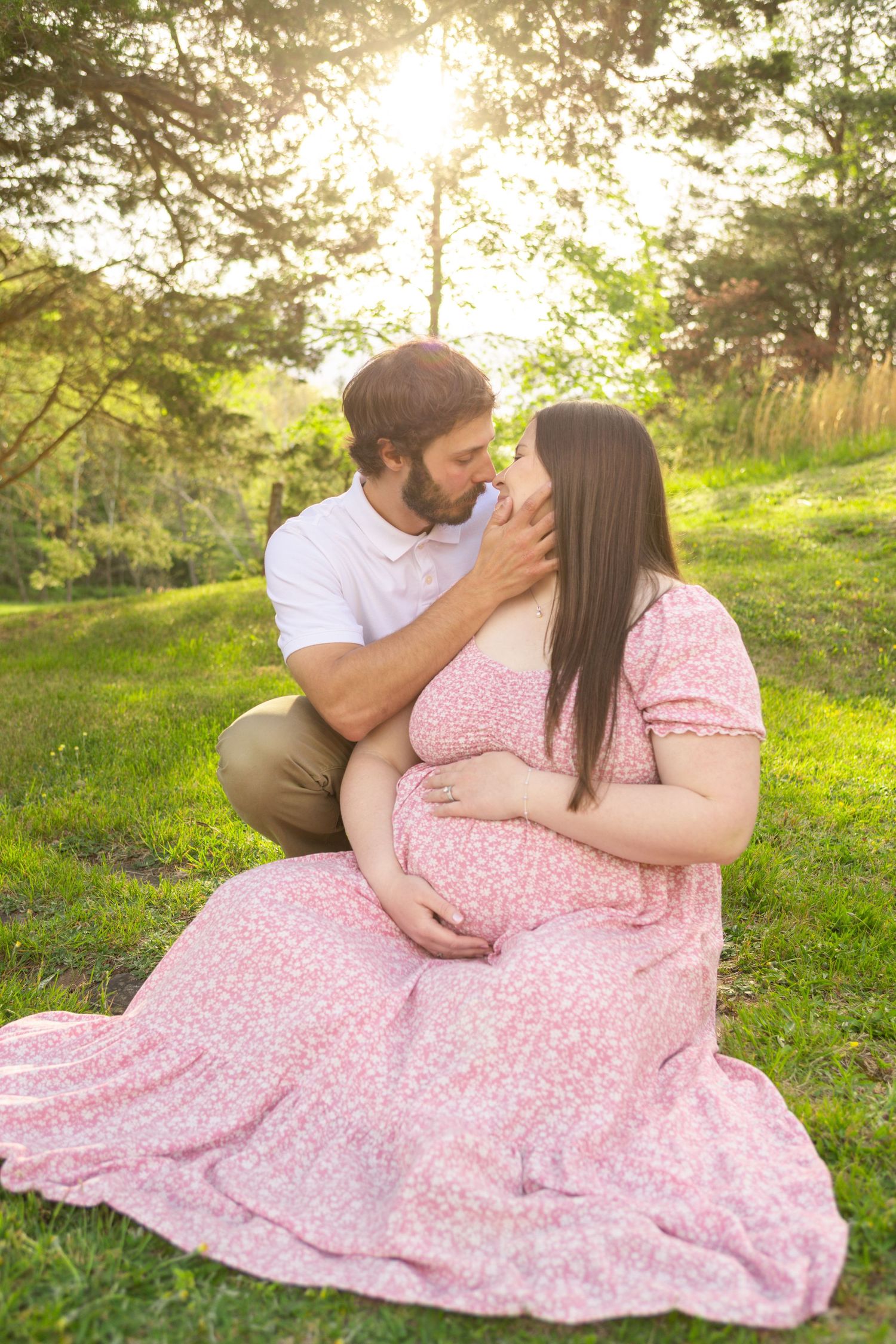 Pregnant couple sharing a tender moment outdoors, woman in pink floral dress cradling baby bump, golden sunlight filtering through trees.