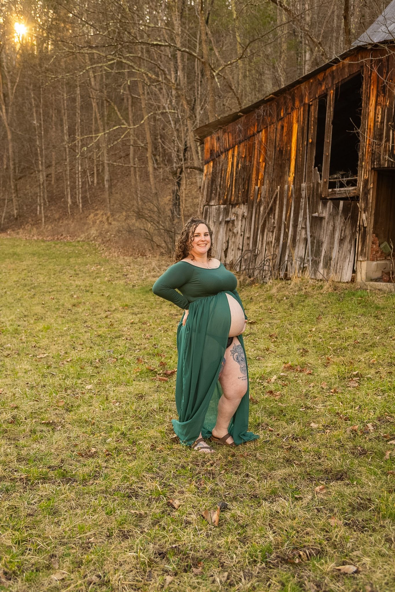 Pregnant woman in green sheer maternity gown posing at golden hour near a rustic barn surrounded by bare trees.