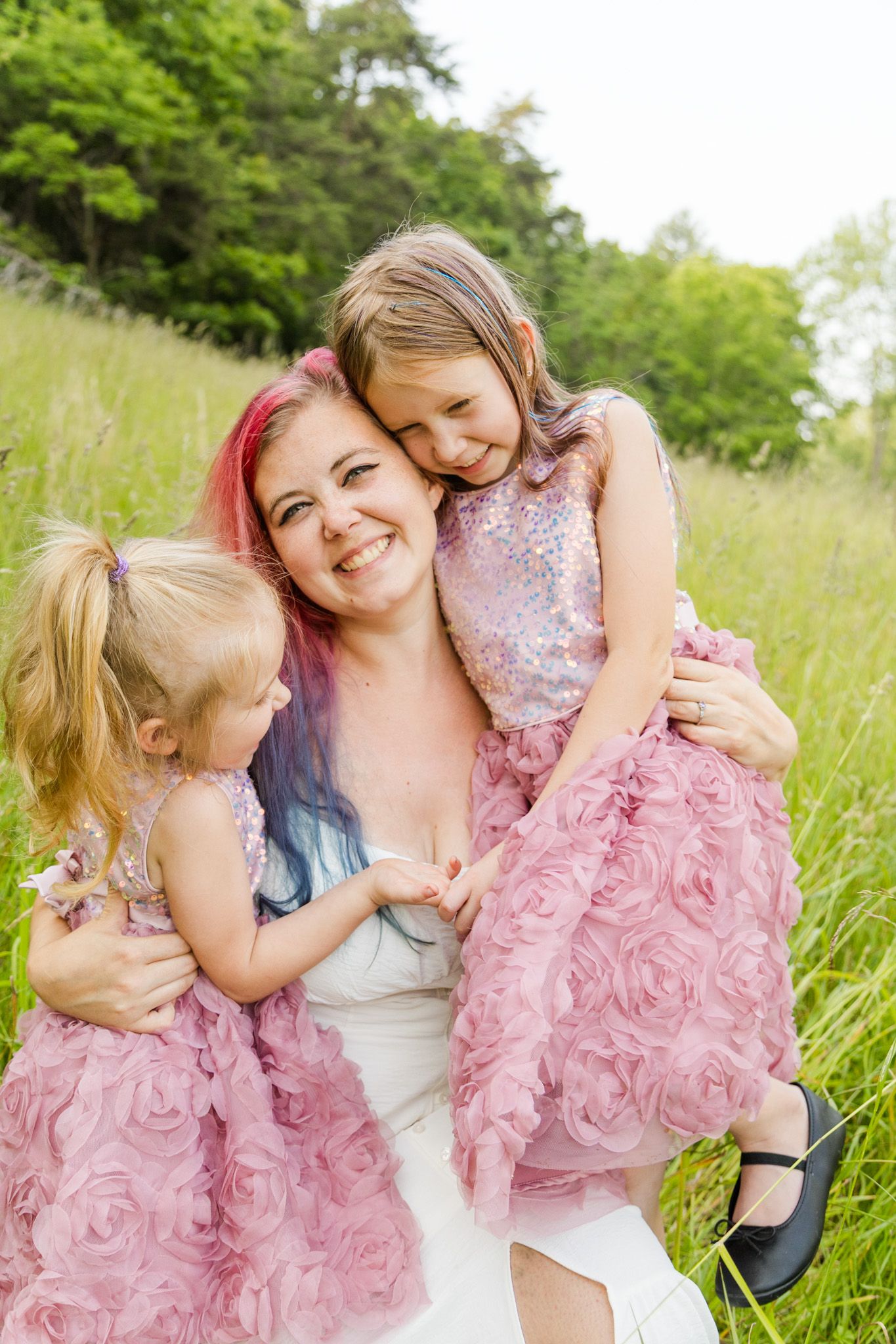 Happy mother with colorful hair hugging two young daughters in matching pink rose dresses in a green meadow.
