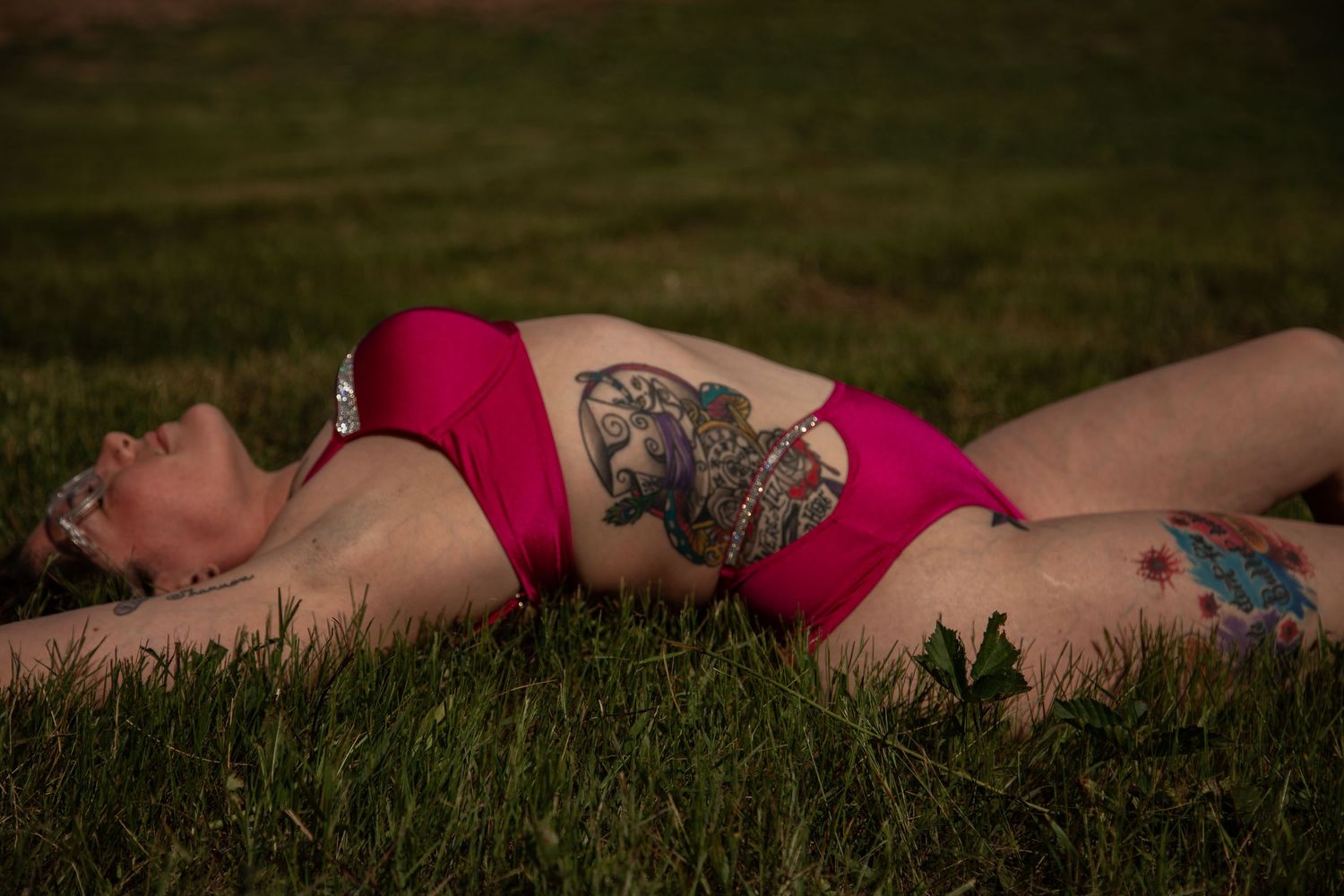 Woman with tattoos lying in grass wearing a pink bikini on a warm evening outdoors.