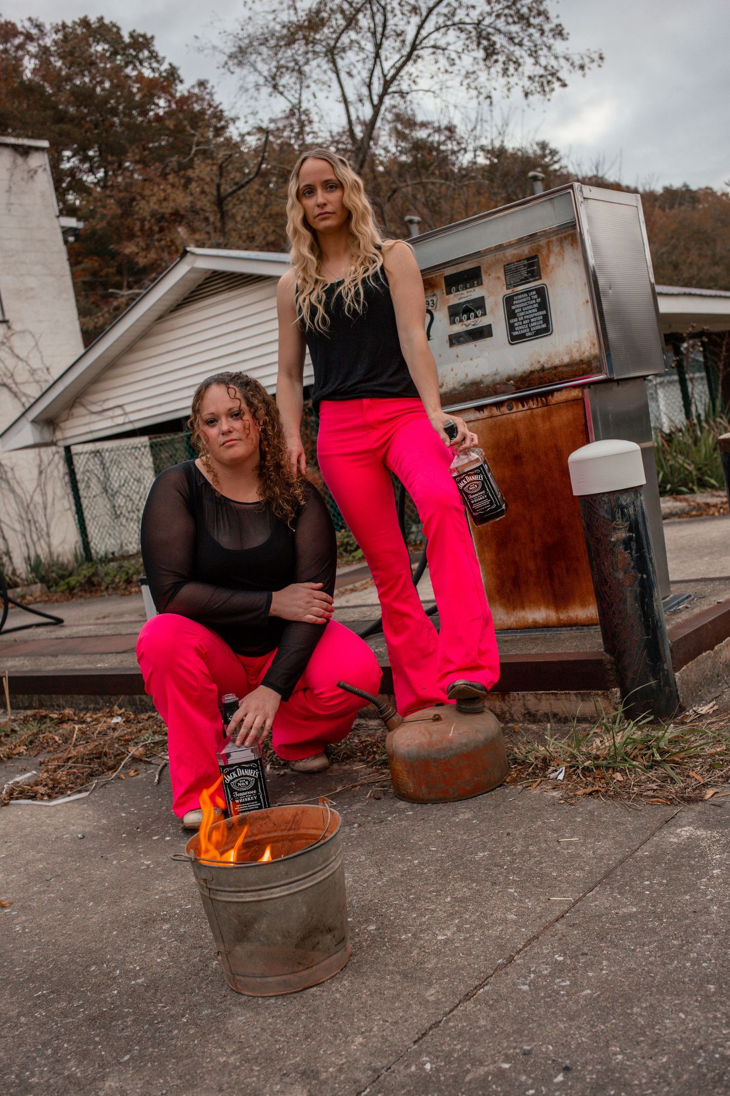 Two women in matching pink flared pants posing with Jack Daniels bottles near a fire barrel outdoors.