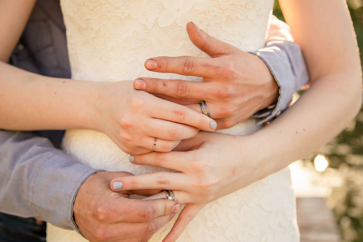 Close-up of newlyweds hands showing wedding rings, bride in white dress and groom in gray suit embracing.