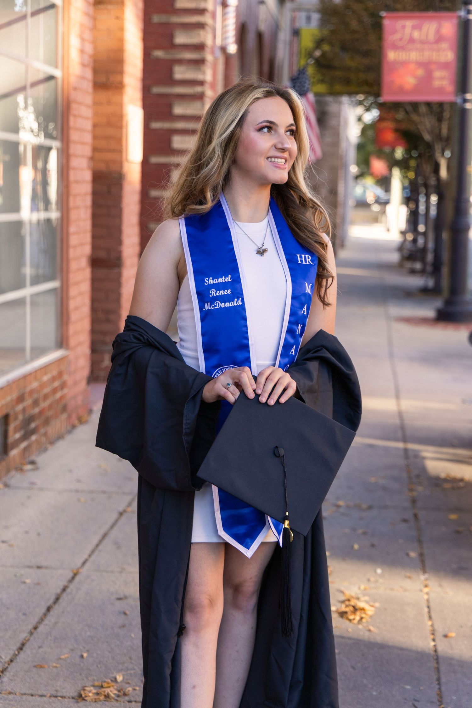 Smiling graduate in a white dress and blue personalized stole holds a black mortarboard while wearing an open gown on a brick-lined street.