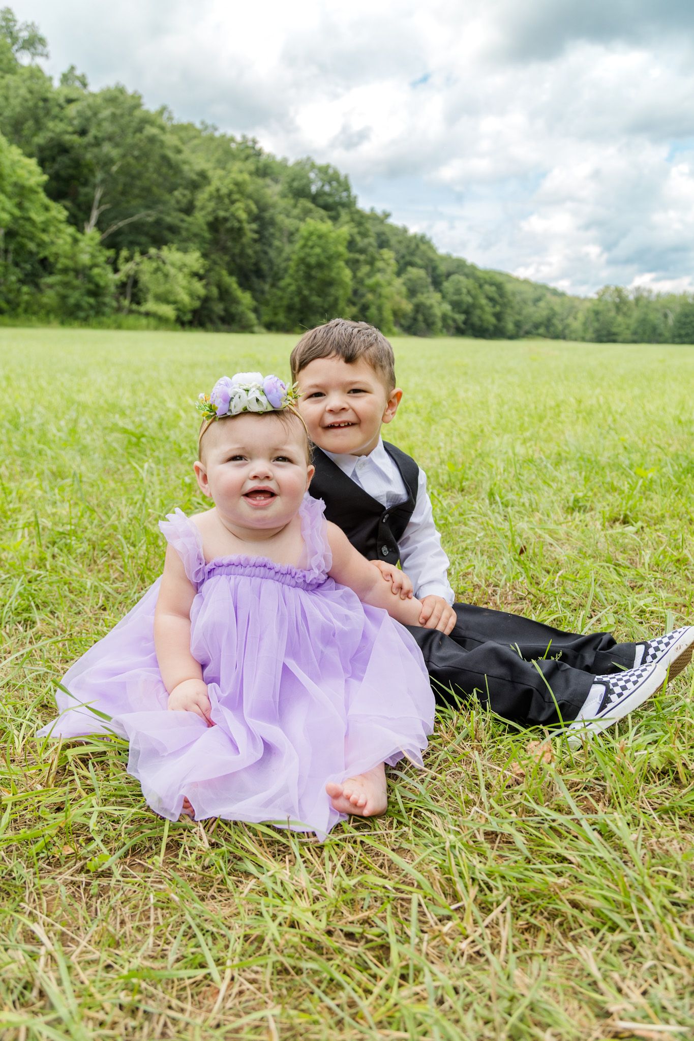 Smiling baby girl in purple dress and toddler boy in tuxedo sitting together on a green grassy field.