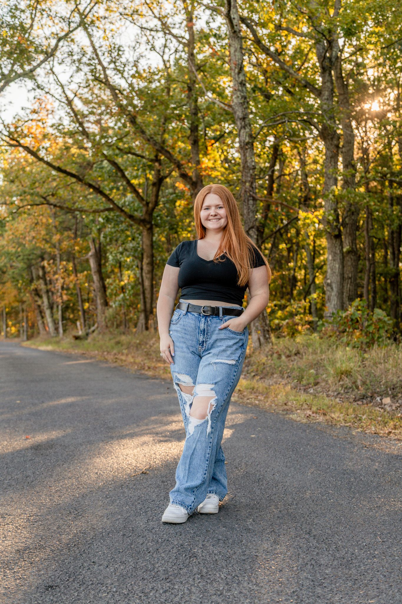 Smiling redhead teen in black top and ripped jeans standing on a tree-lined road at sunset.