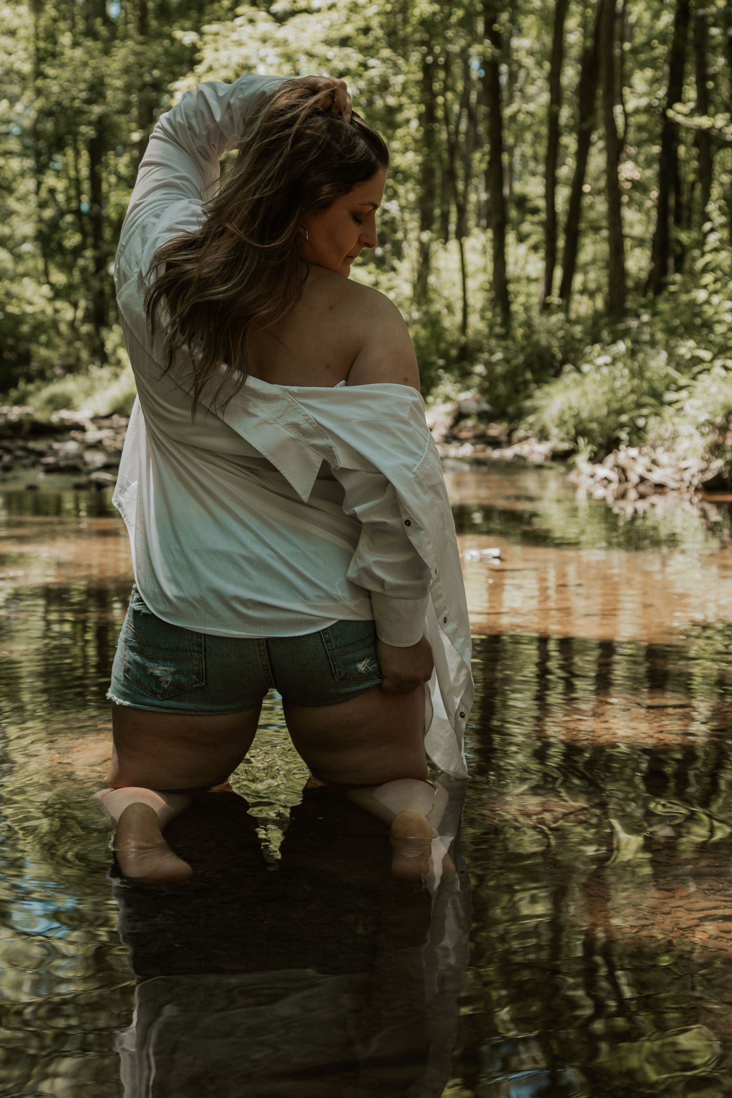 Woman in white off-shoulder shirt and denim shorts wading in a shallow forest creek.