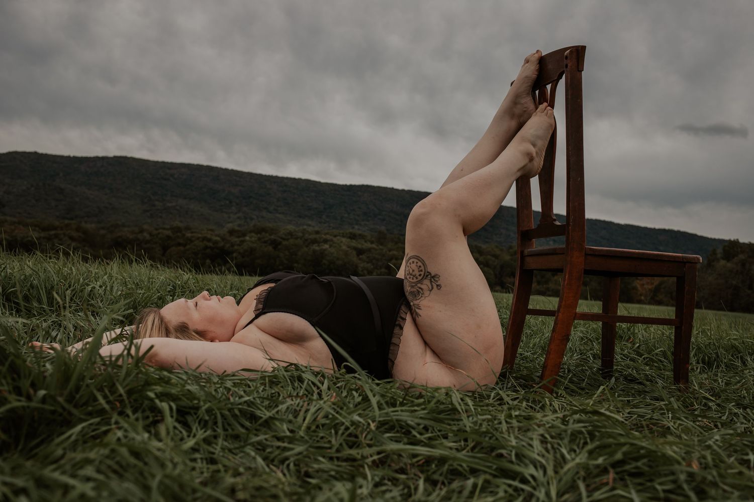 Blonde woman in black bodysuit lying in grass with legs resting on a wooden chair outdoors.