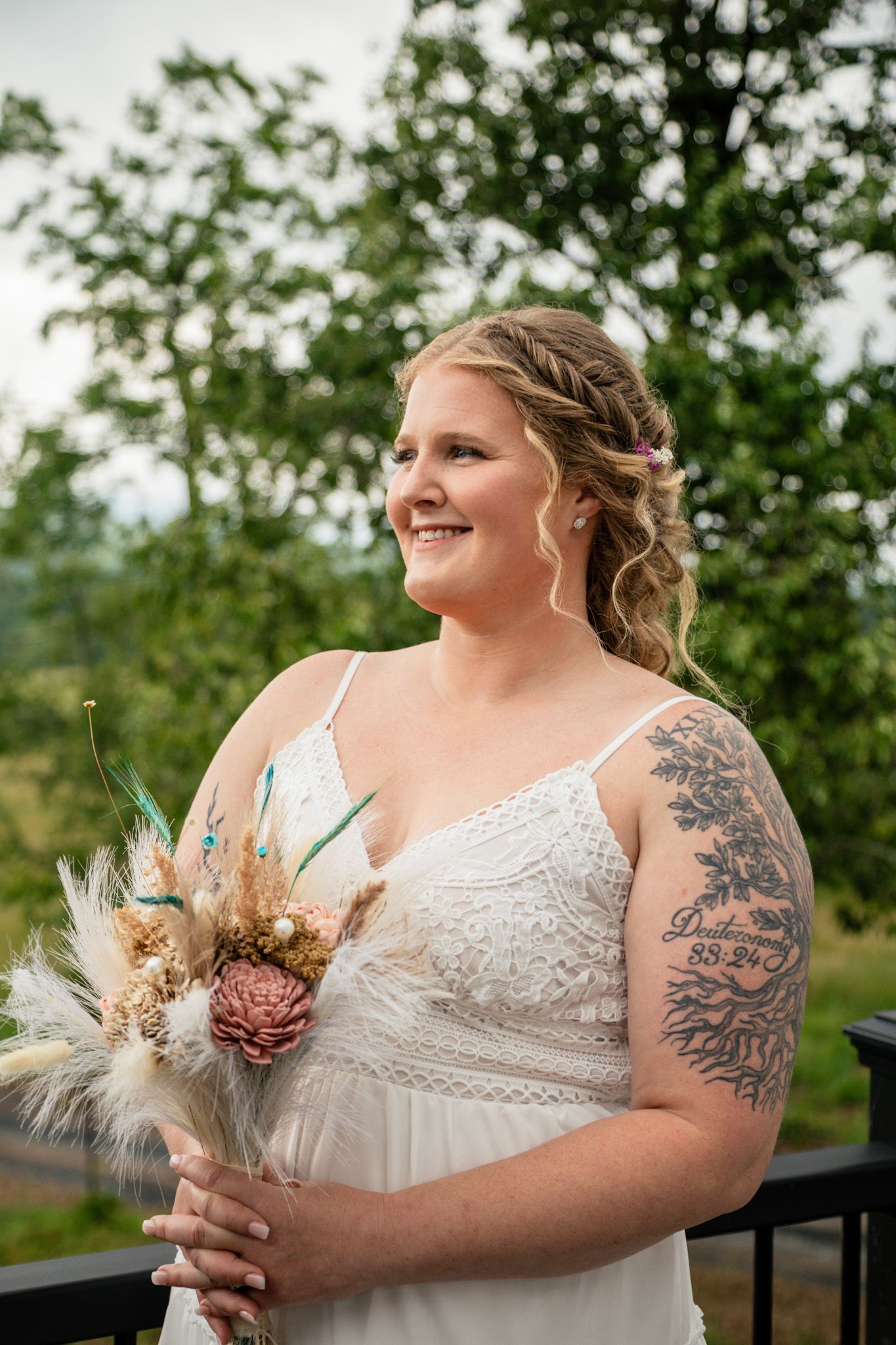 Smiling bride with braided hair and arm tattoo holding boho dried flower bouquet outdoors.