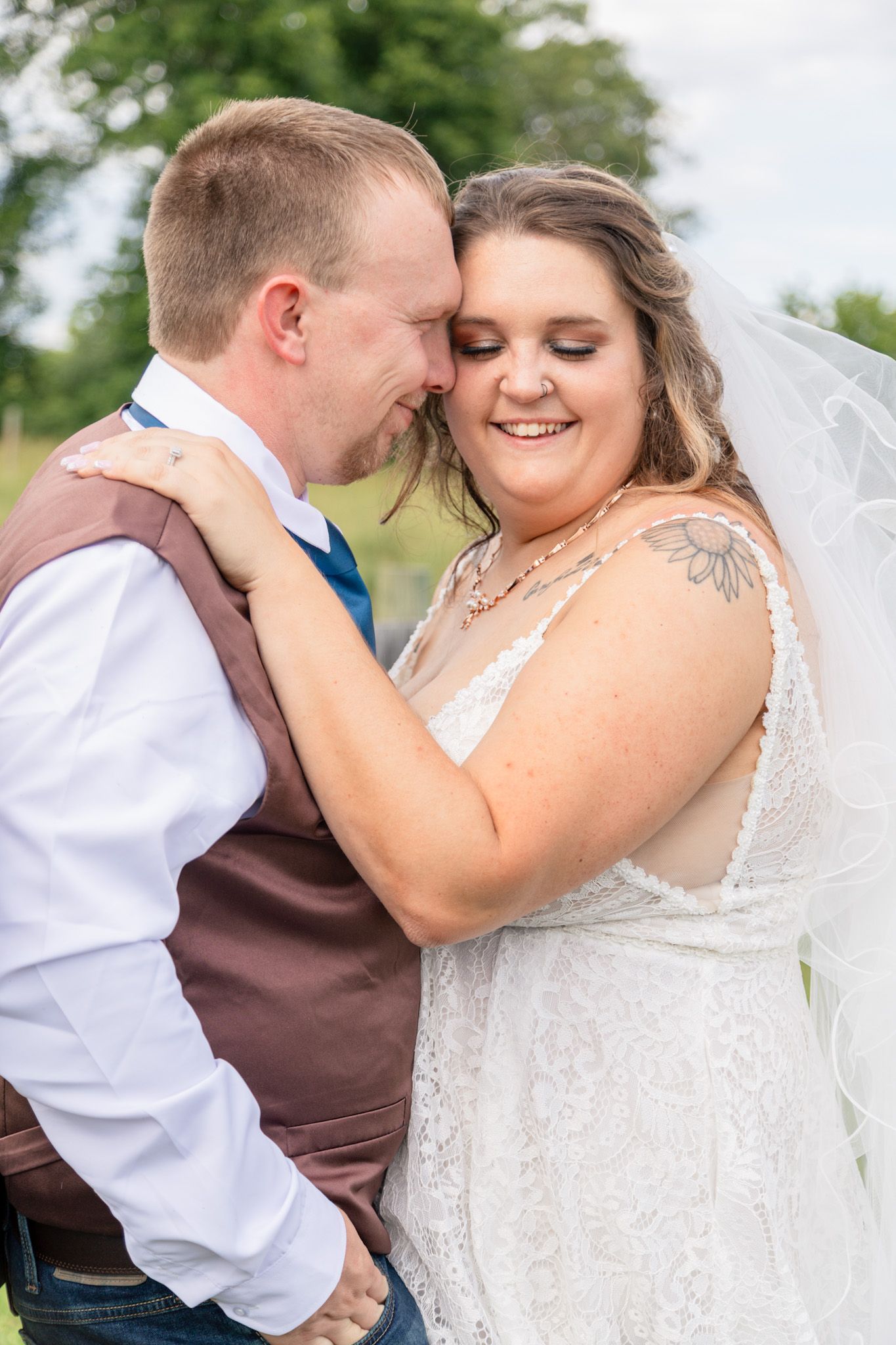 Smiling bride with veil embracing groom in a vest outdoors at a farm wedding.