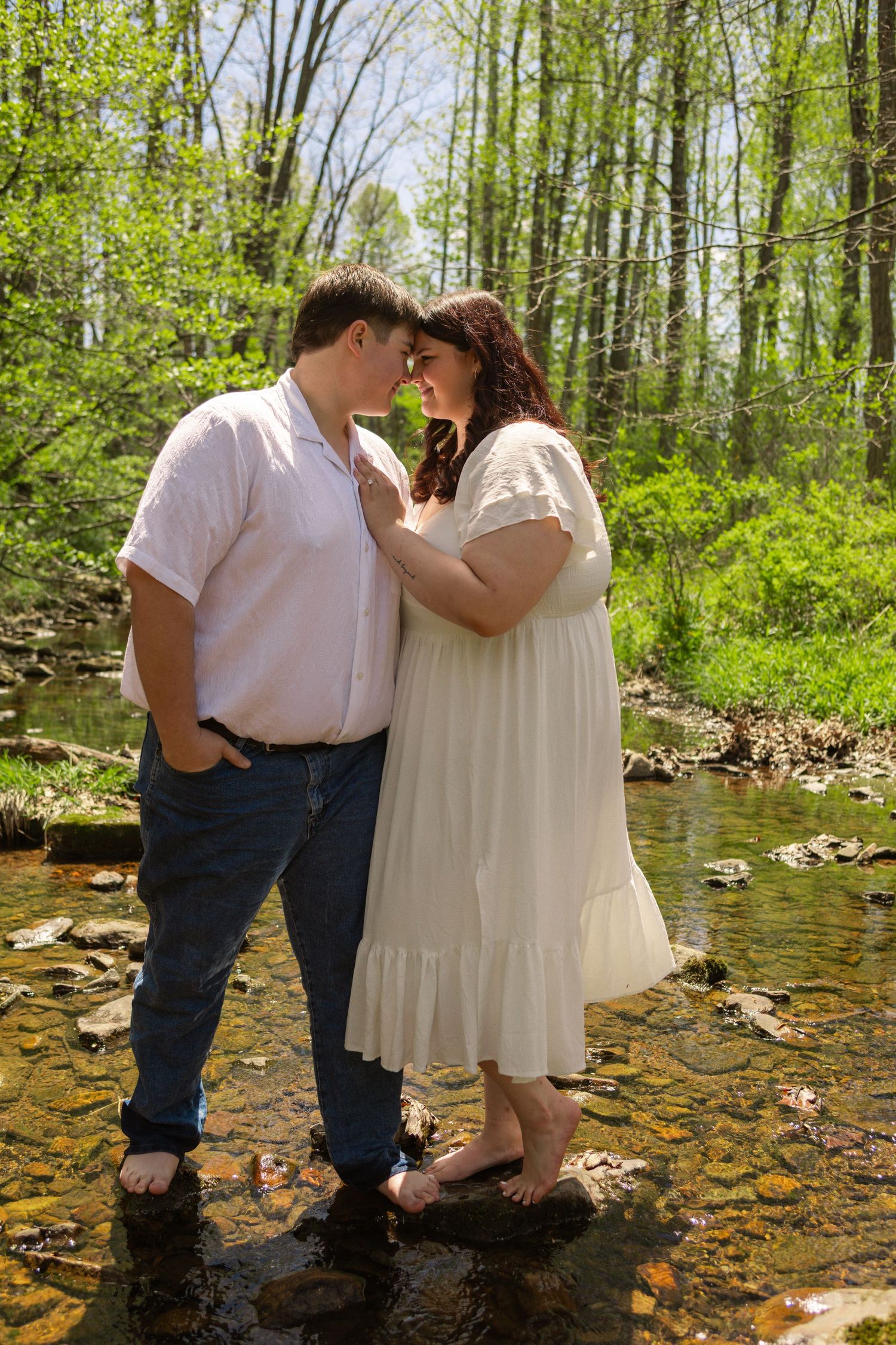 Couple standing barefoot in a rocky creek, surrounded by lush green forest, sharing a romantic moment.