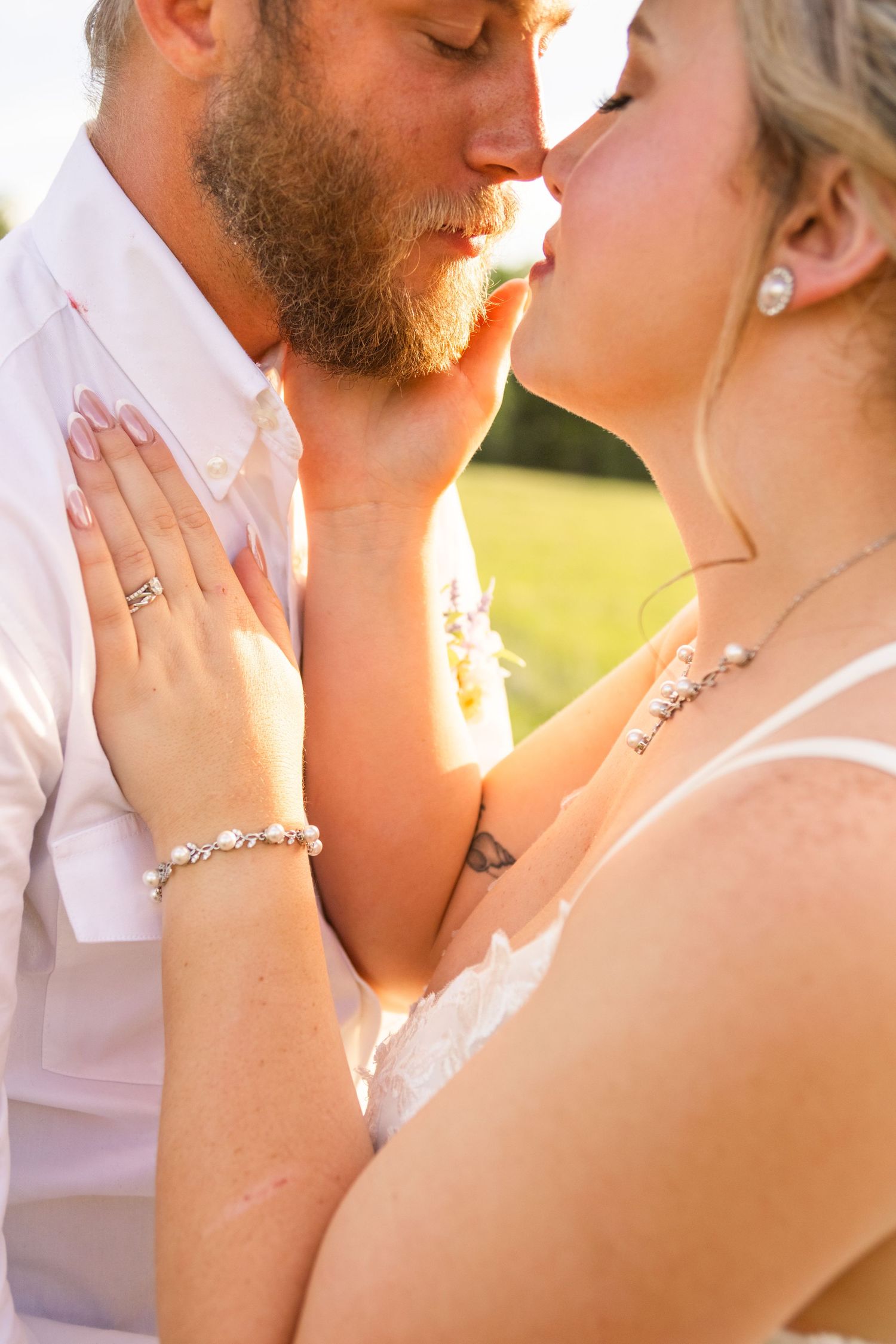 A bride and groom share a romantic kiss outdoors, with the bride wearing a diamond bracelet and wedding ring in golden sunlight.