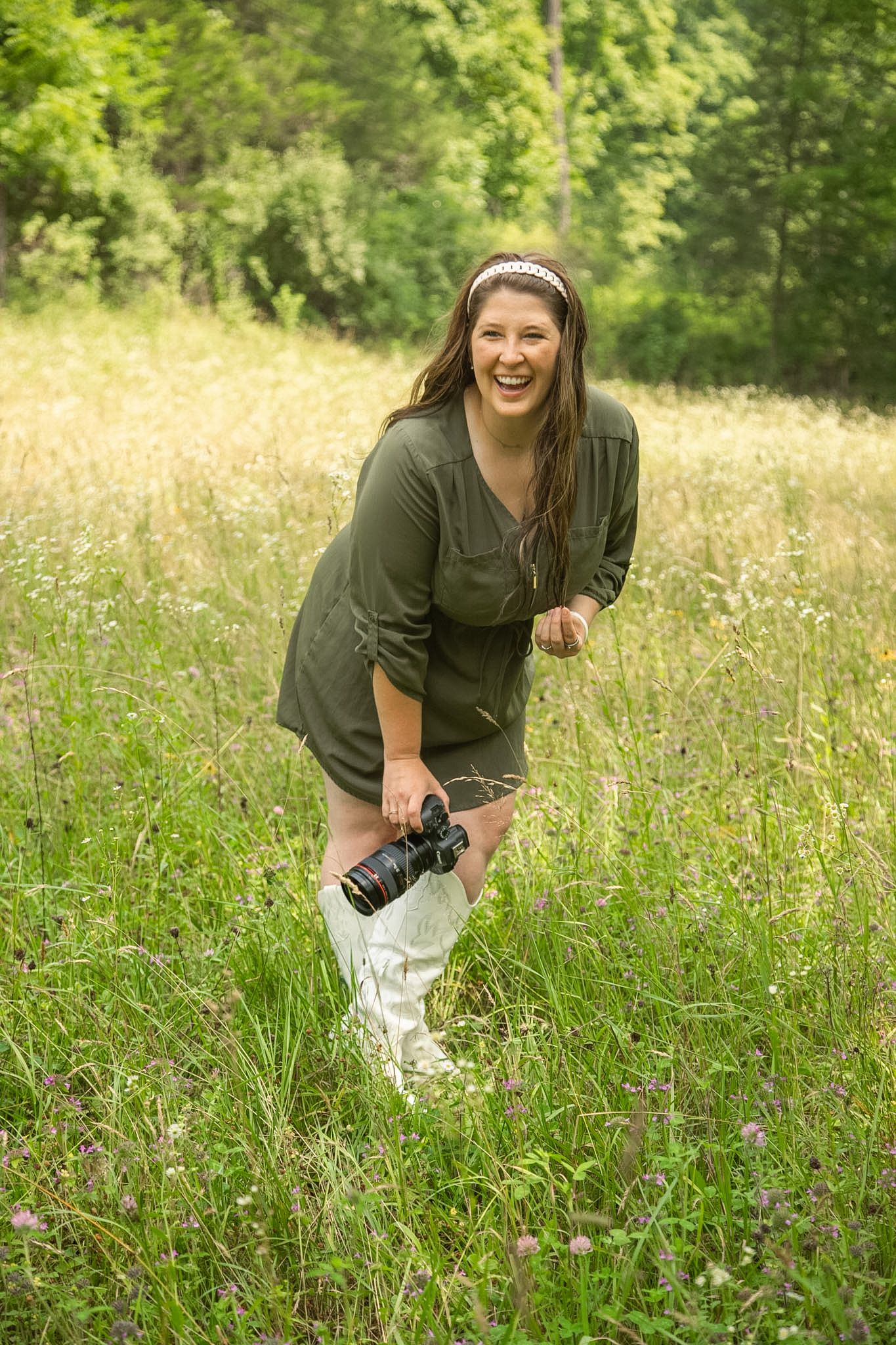 A smiling woman in a green dress holds a DSLR camera while standing in a lush meadow surrounded by wildflowers.
