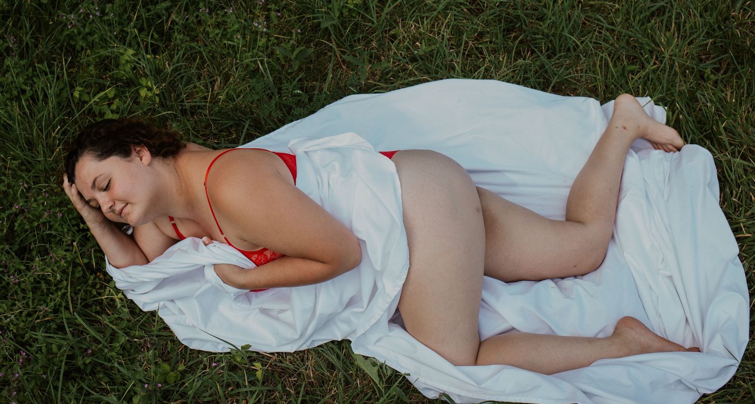 Pregnant woman in red bikini resting on white fabric in green grass outdoors.