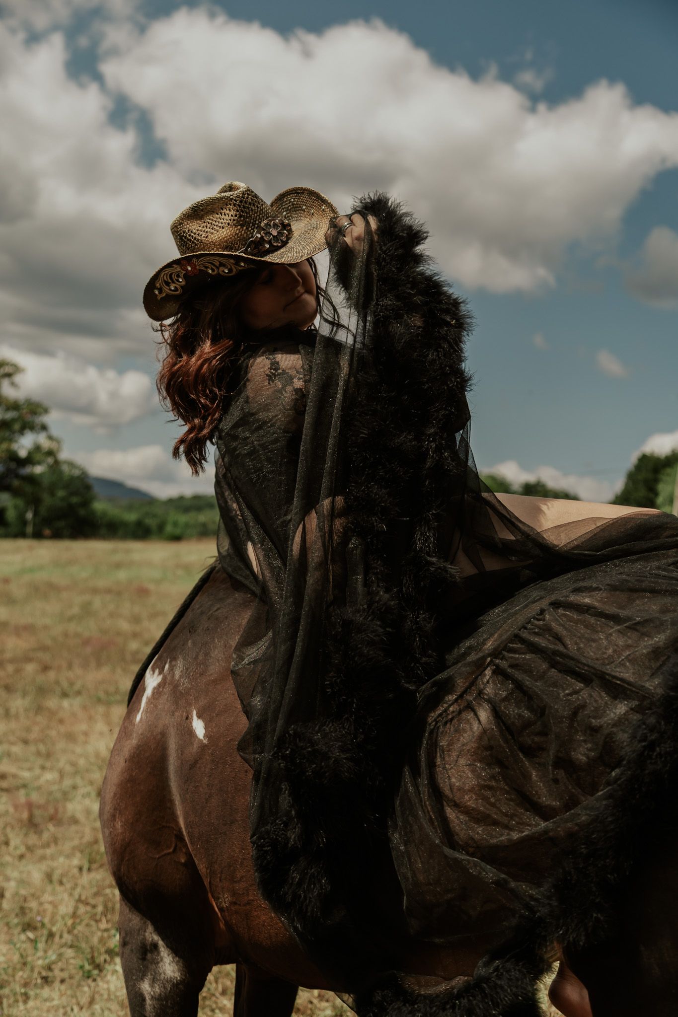 Woman in cowboy hat and black lace cape riding a brown horse in an open field under cloudy skies.