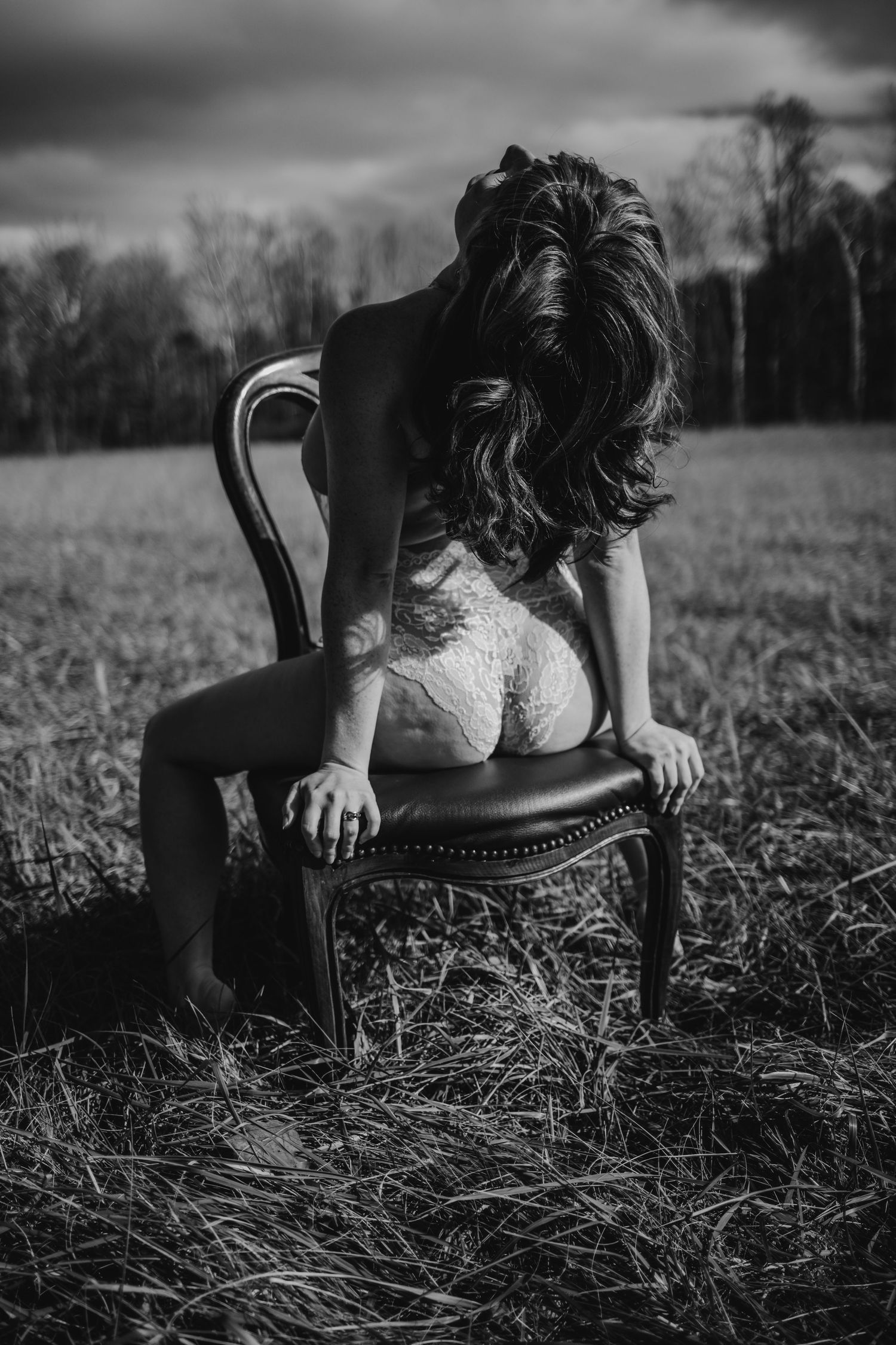 Black and white photo of a woman with long hair sitting on a vintage chair in an open field.