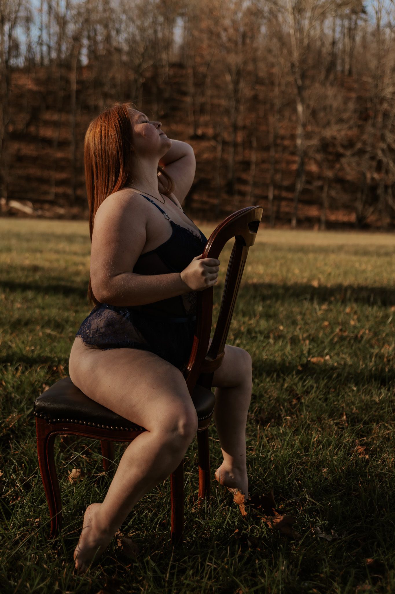 Redhead woman in black lace bodysuit poses on vintage chair outdoors in warm autumn light.