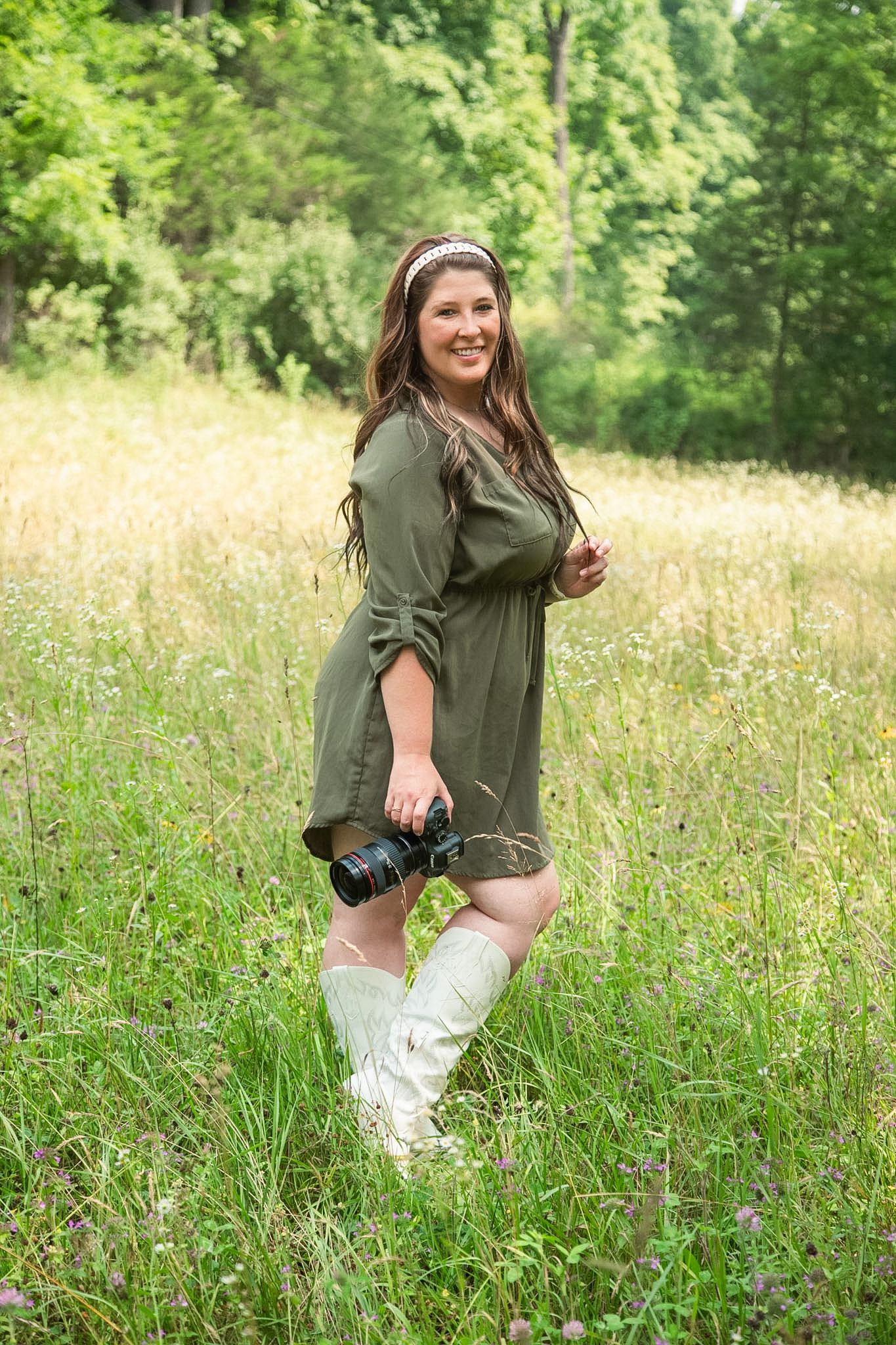 Smiling woman in green dress and white boots holding a camera in a lush green meadow.