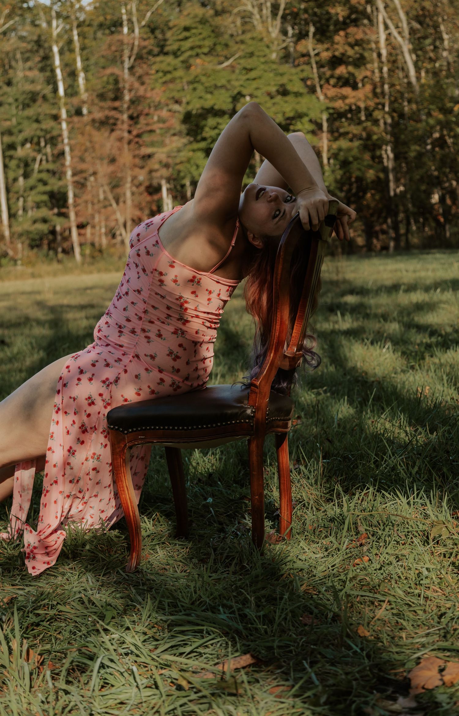 A woman in a pink floral dress poses beside a vintage chair in a sunlit forest clearing.