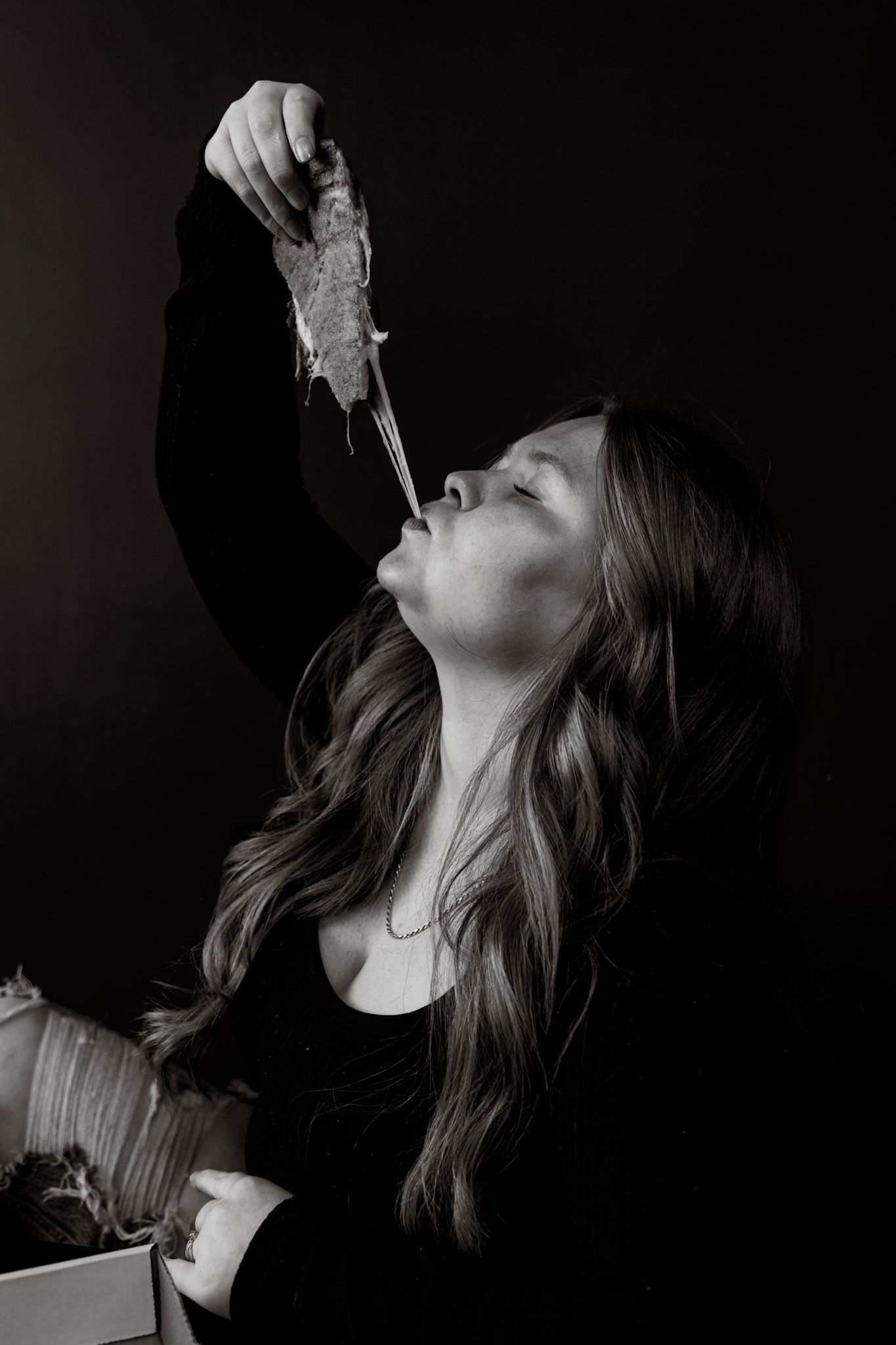 Black and white portrait of a long-haired woman tilting her head back holding food above her mouth.