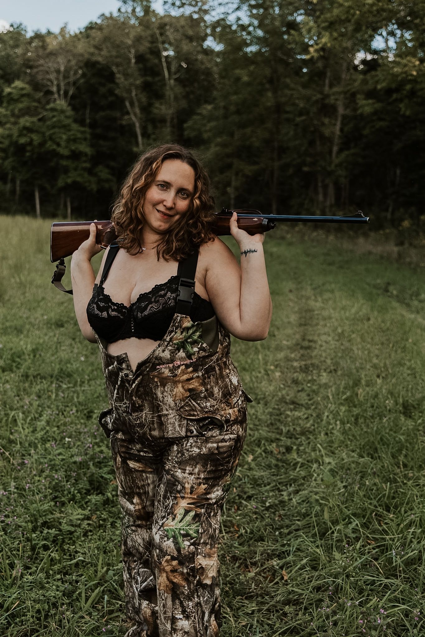 Woman with curly hair smiles in a grassy field wearing camouflage overalls and a black lace top, with a rifle resting on her shoulders.