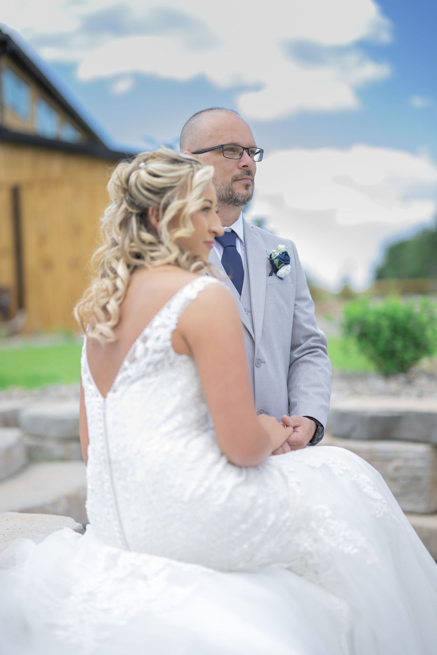 Bride in white lace gown and groom in gray suit sit together outdoors at a sunny wedding venue.